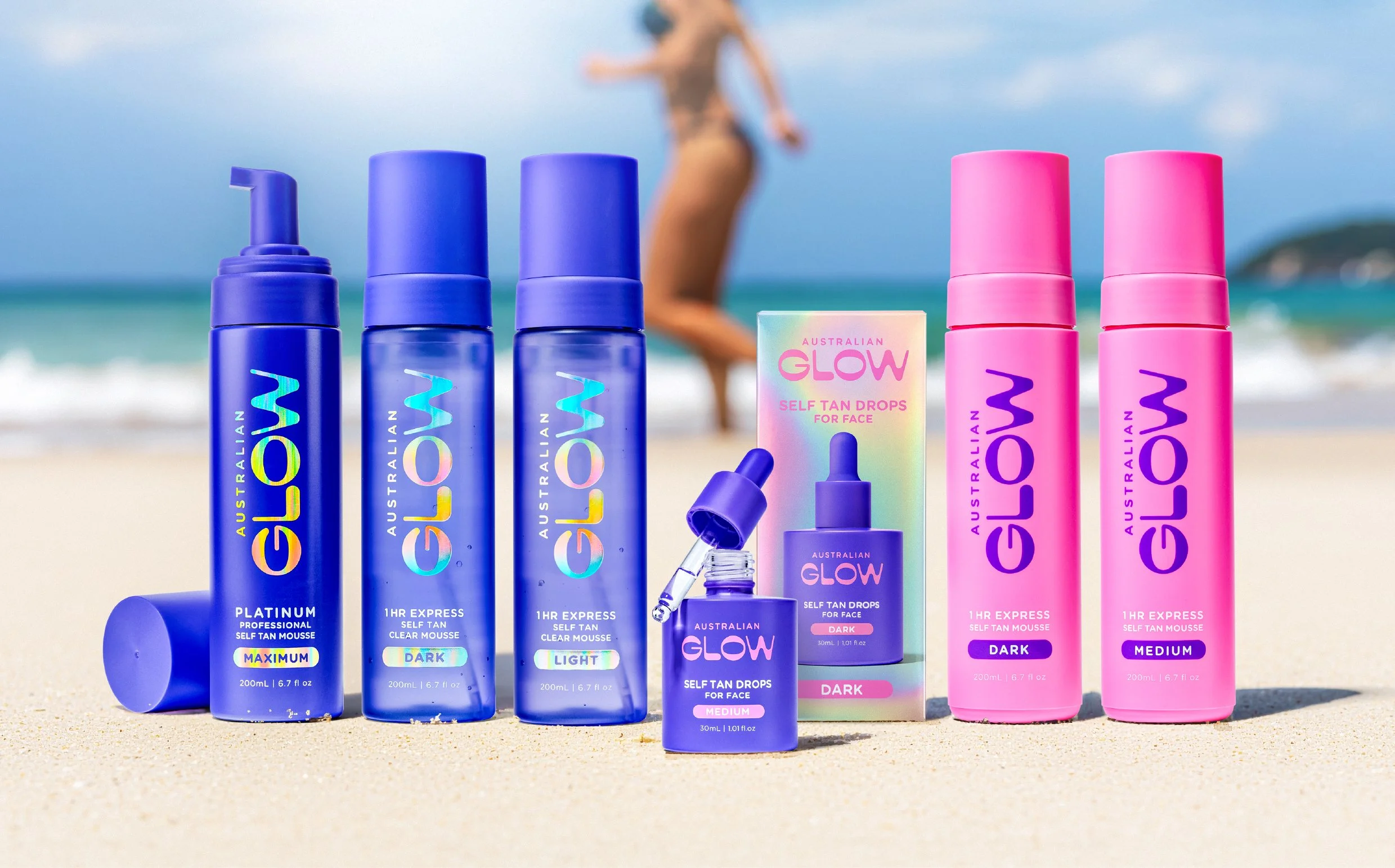 Collection of Australian Glow self-tan products on a beach, including mousse bottles in blue and pink, with a blurred person in swimwear in the background.