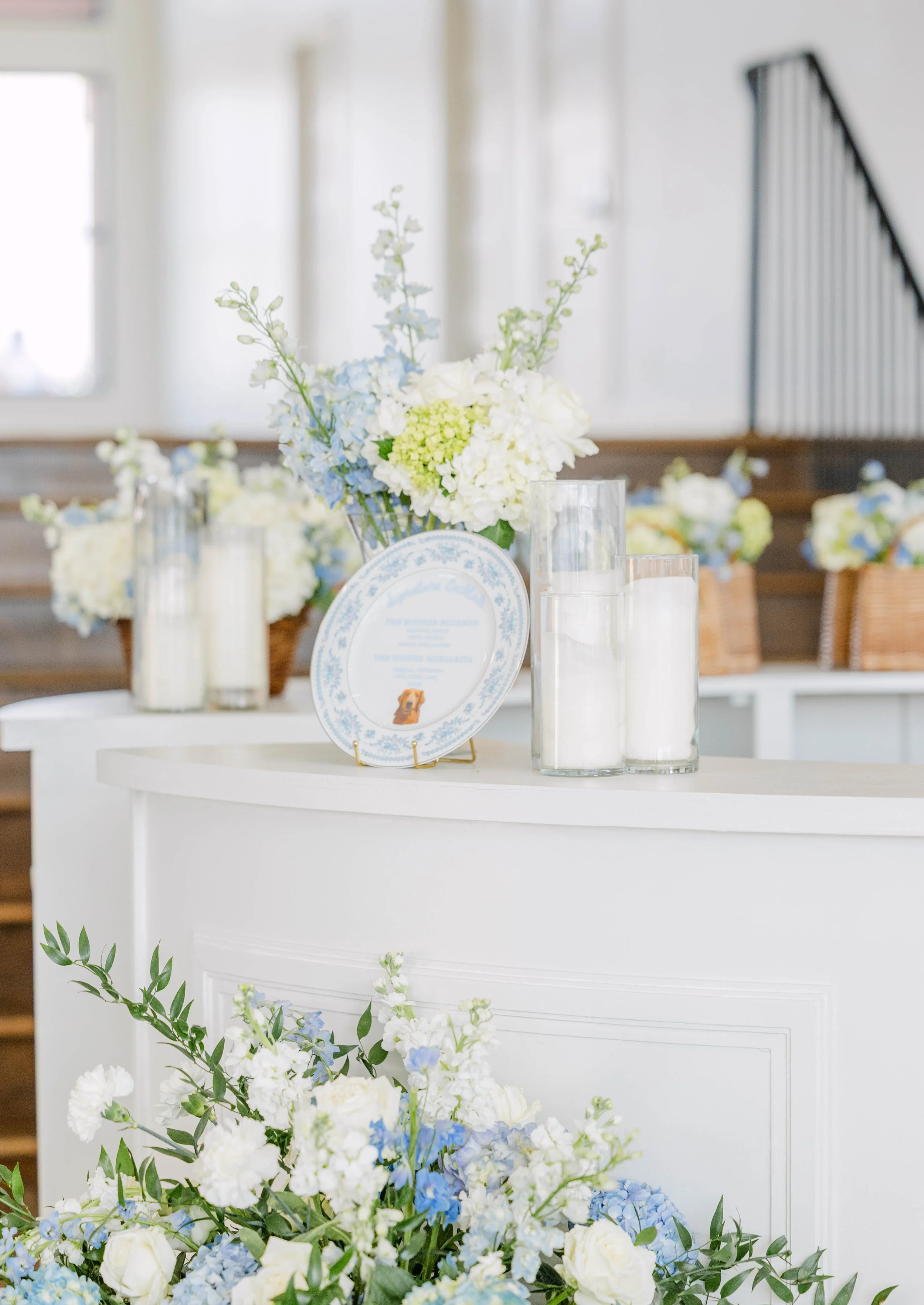 A decorated table with a bouquet of white and blue flowers, candles, and a framed note, set in a room with wooden stairs and natural light.