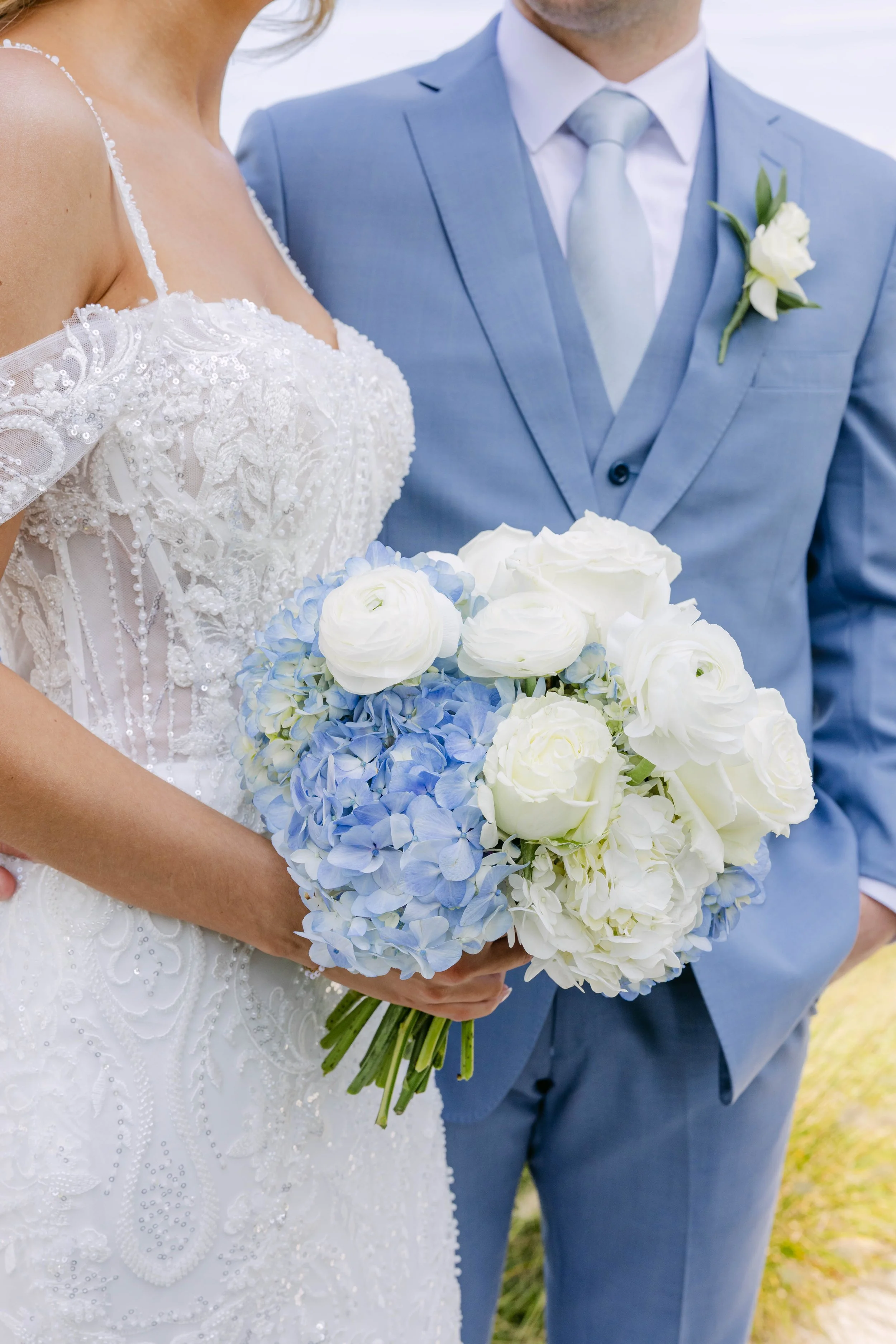 Close-up of a bride holding a bouquet of white roses and blue hydrangeas, with a groom standing next to her in a light blue suit and a white boutonniere.