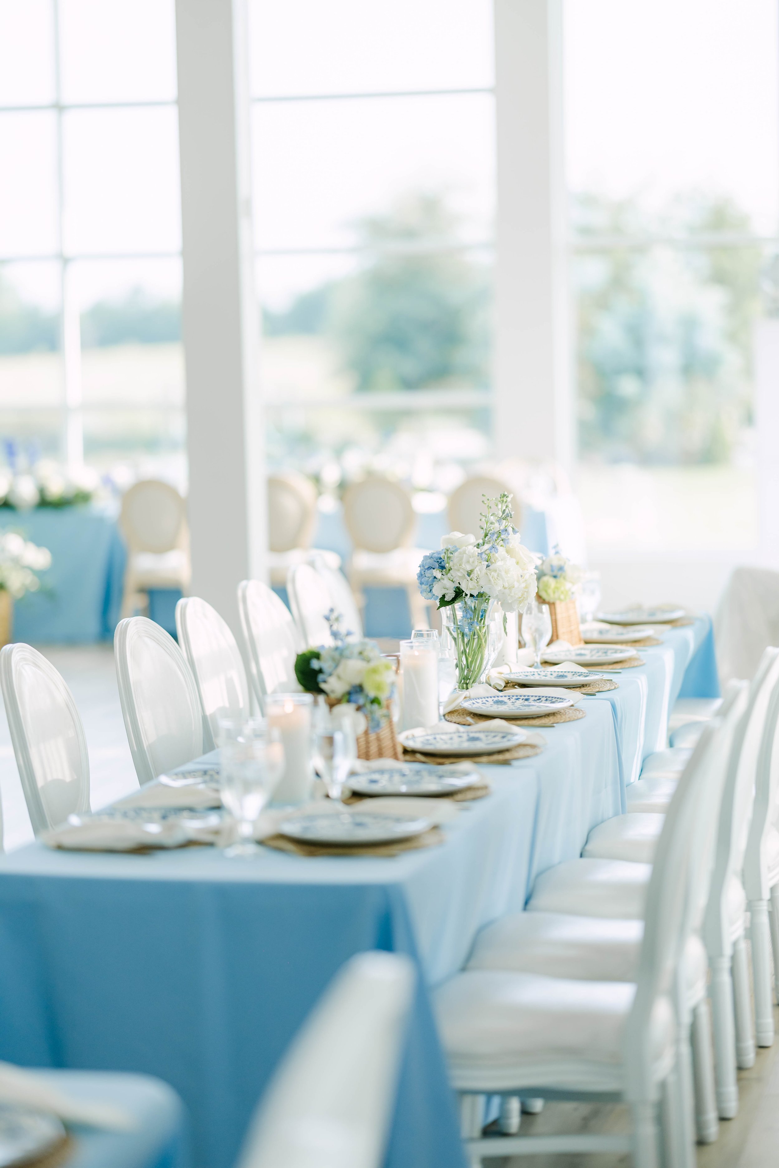 Elegant banquet table decorated with blue and white floral arrangements, candles, and china, set up for a celebration in a bright, airy room with large windows.