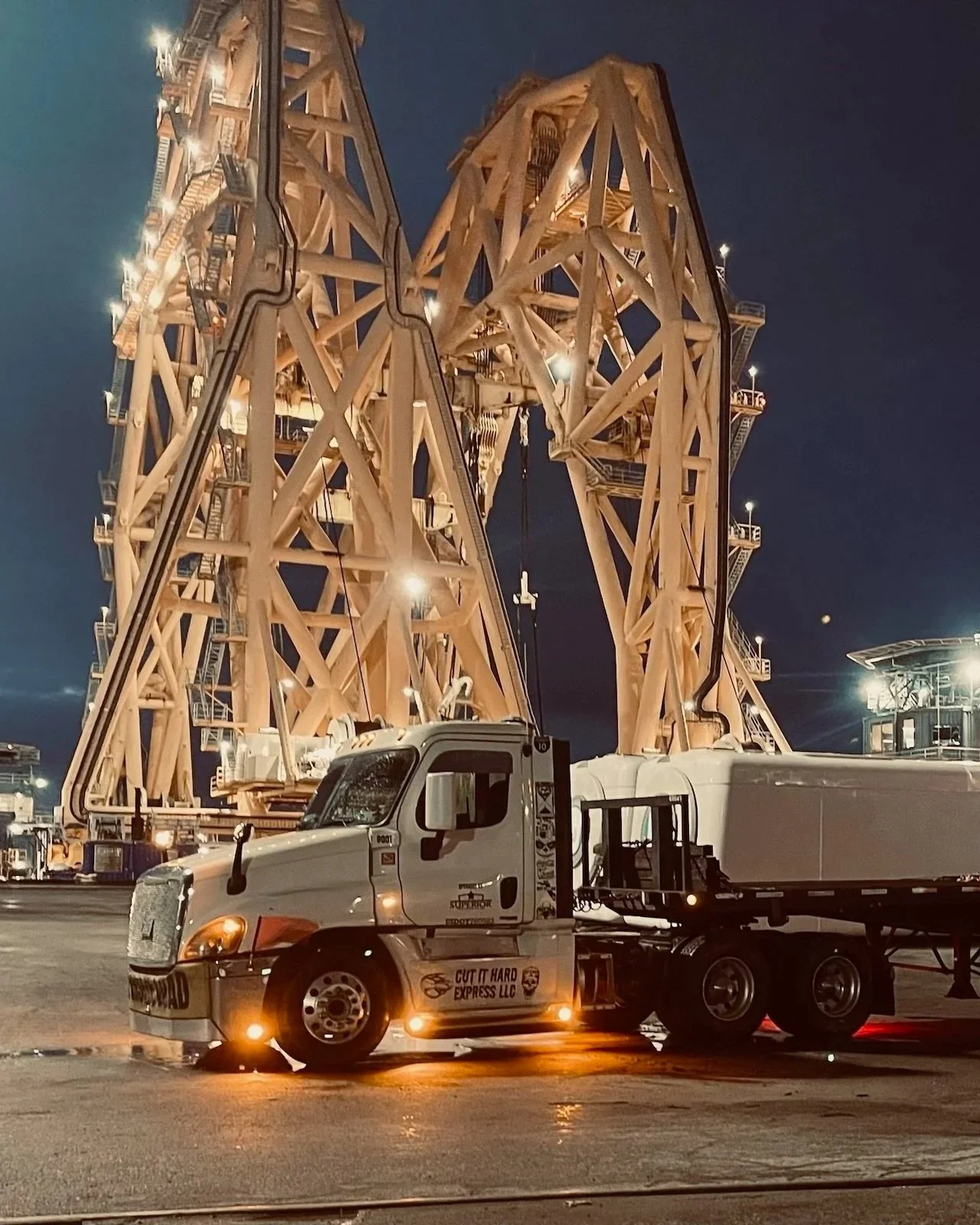 Nighttime scene of a large wooden amusement park ride, perhaps a drop tower, with bright lights. A white truck labeled 'Cut It Hard Express LLC' is parked in front of it, with small orange lights under the truck.