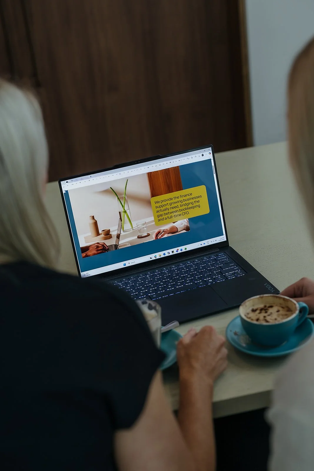 Two women seated at a table, looking at a laptop screen with a presentation slide. One woman has blonde hair, and the other a darker complexion. There are two cups of coffee on the table, one with a frothy drink and a sprinkle of cocoa or cinnamon.