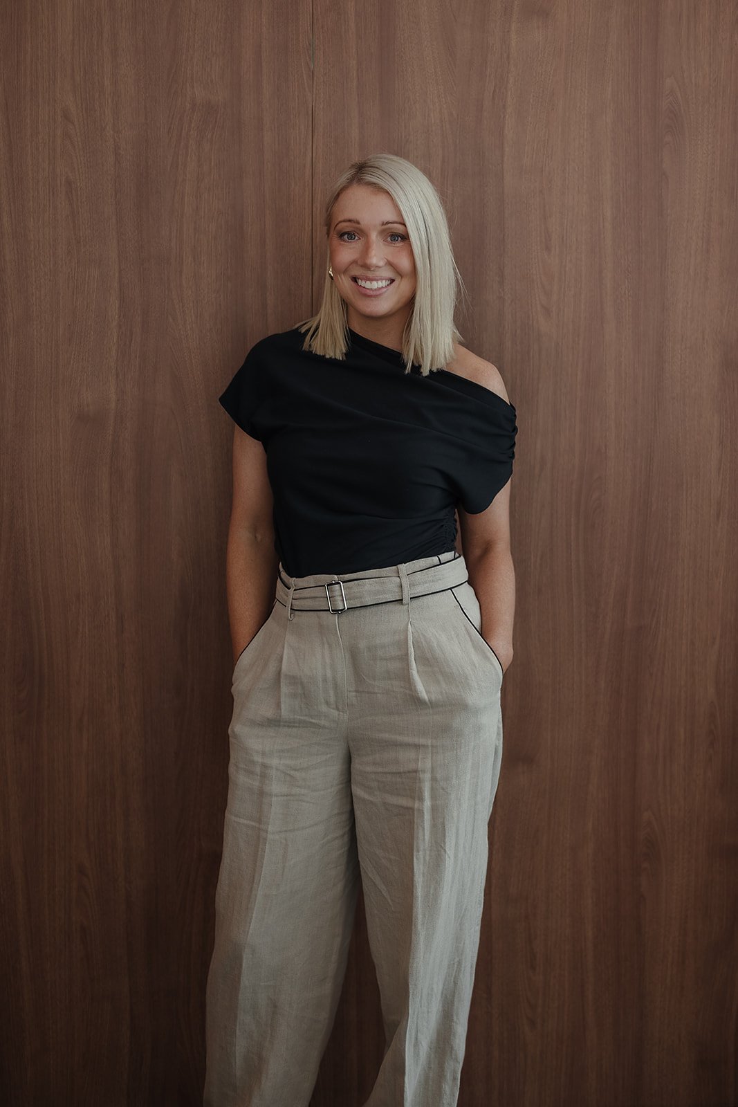 A woman with blonde hair wearing a black, off-the-shoulder top and beige high-waisted trousers standing against a wooden wall.