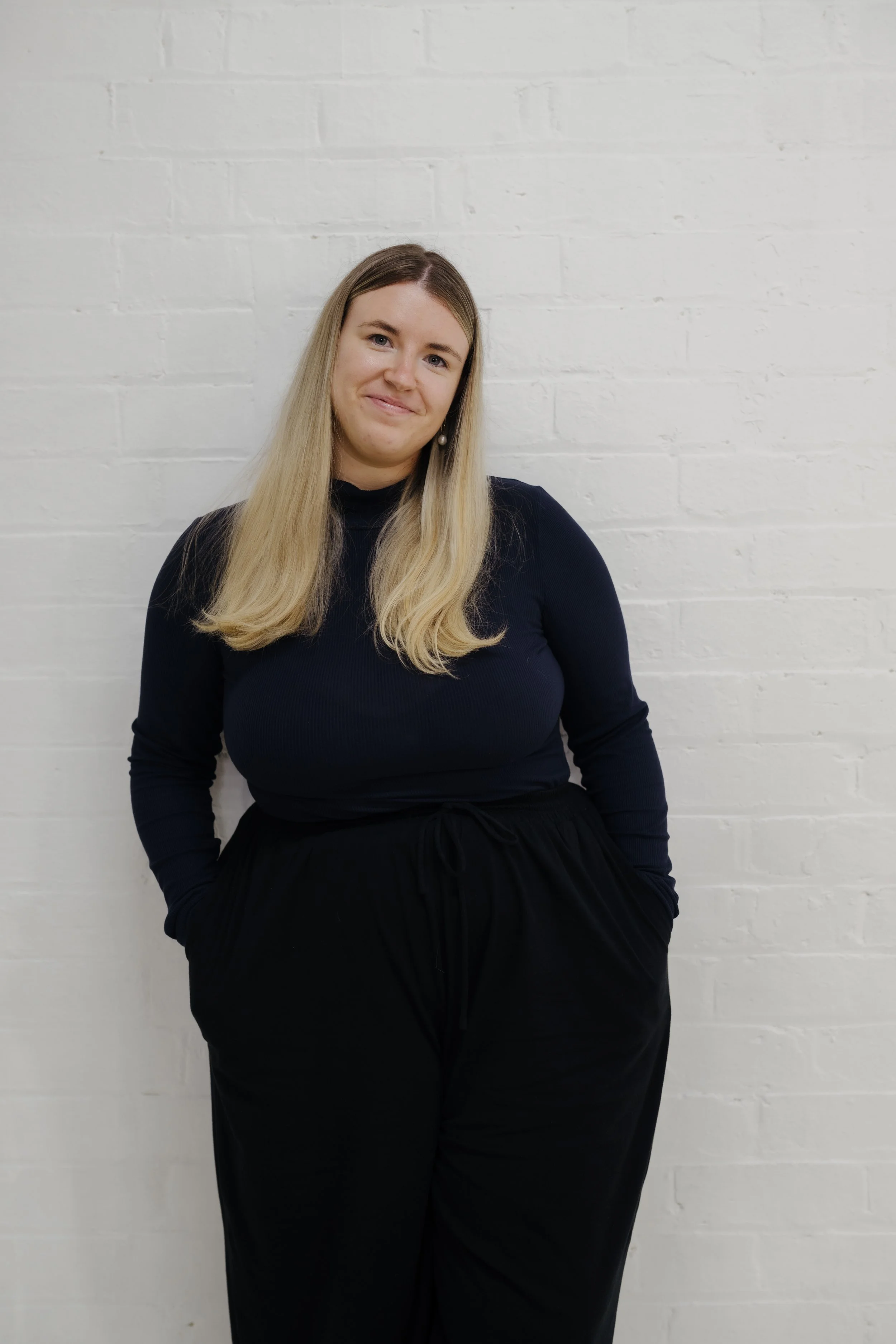 A woman with long blonde hair wearing a dark blue long-sleeve shirt and black pants, standing against a white brick wall, smiling with her hands in her pockets.