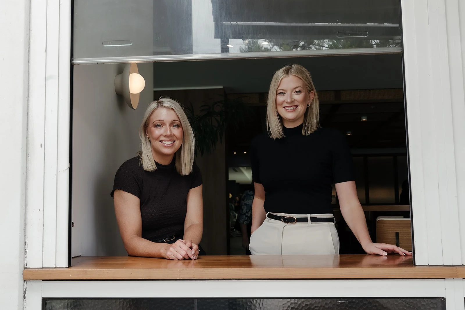 Two women smiling and leaning on a wooden counter at a cafe or restaurant window