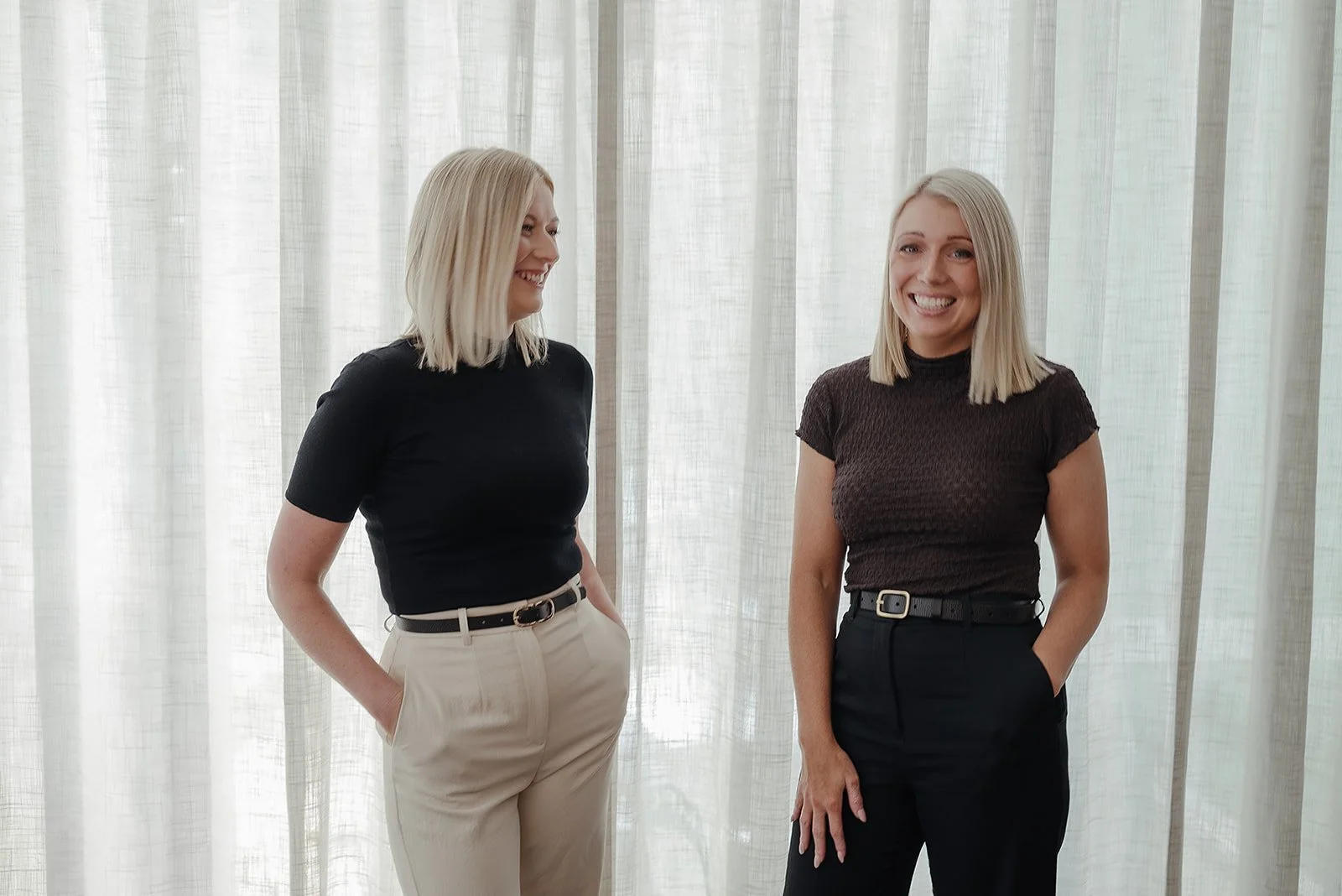 Two women with blonde hair smiling and talking to each other indoors, standing in front of white curtains.