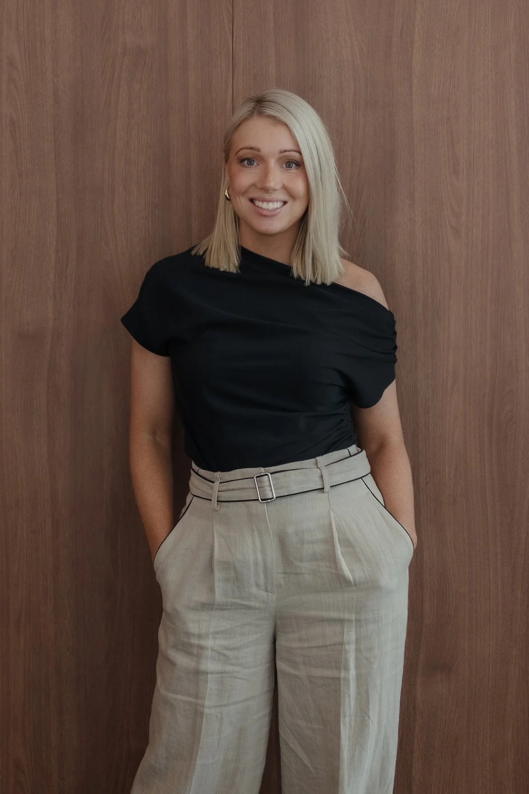 A woman with blonde hair smiling, wearing a black one-shoulder top and beige high-waisted pants, standing in front of a wooden wall.