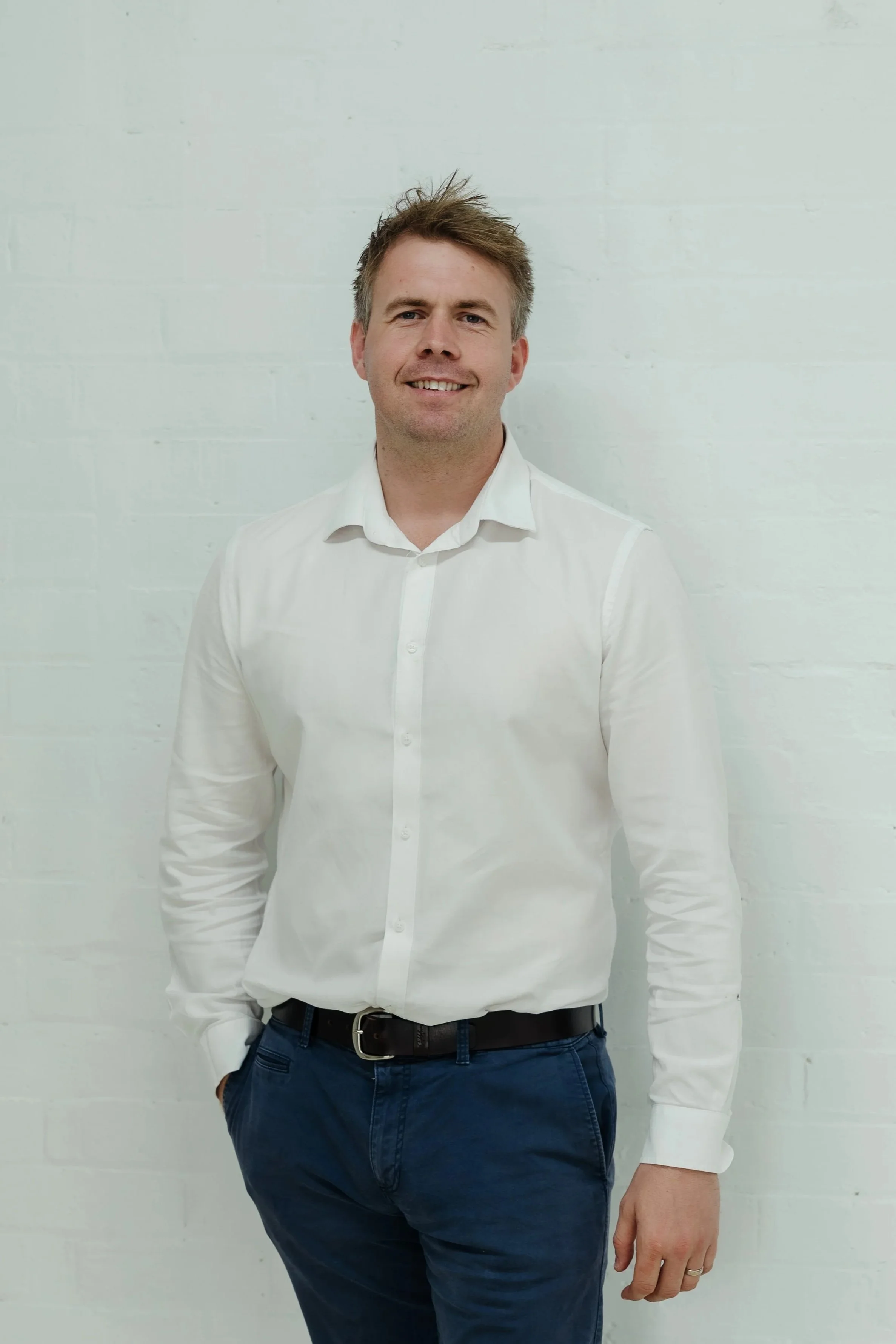 A man with short brown hair, wearing a white shirt and dark blue pants, standing against a white brick wall, smiling with one hand in his pocket.