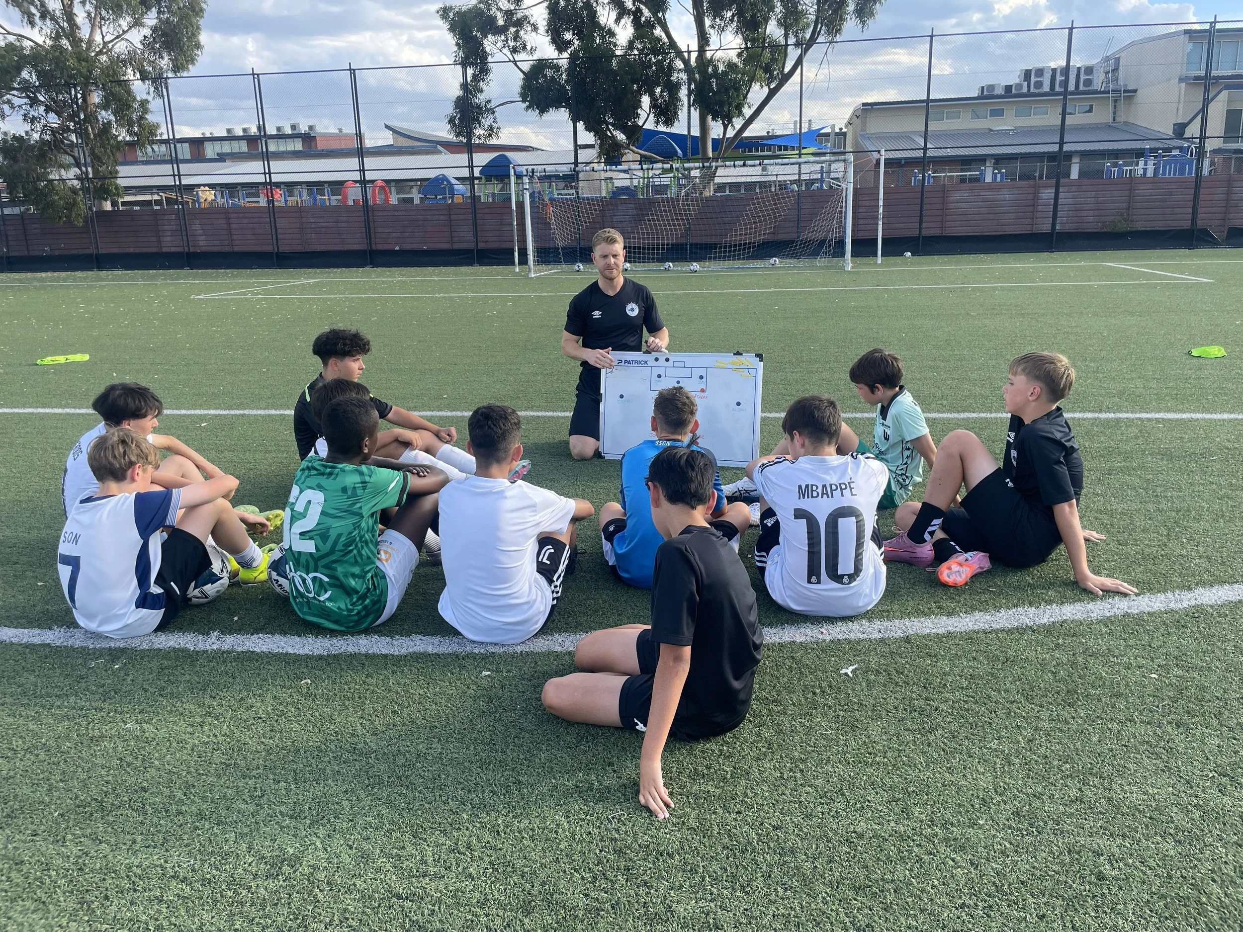Football coach giving instruction to a group of young boys sitting on the field during a practice session.