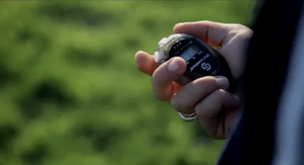 A person holding a small digital stopwatch or timer outdoors with blurred green grass in the background.