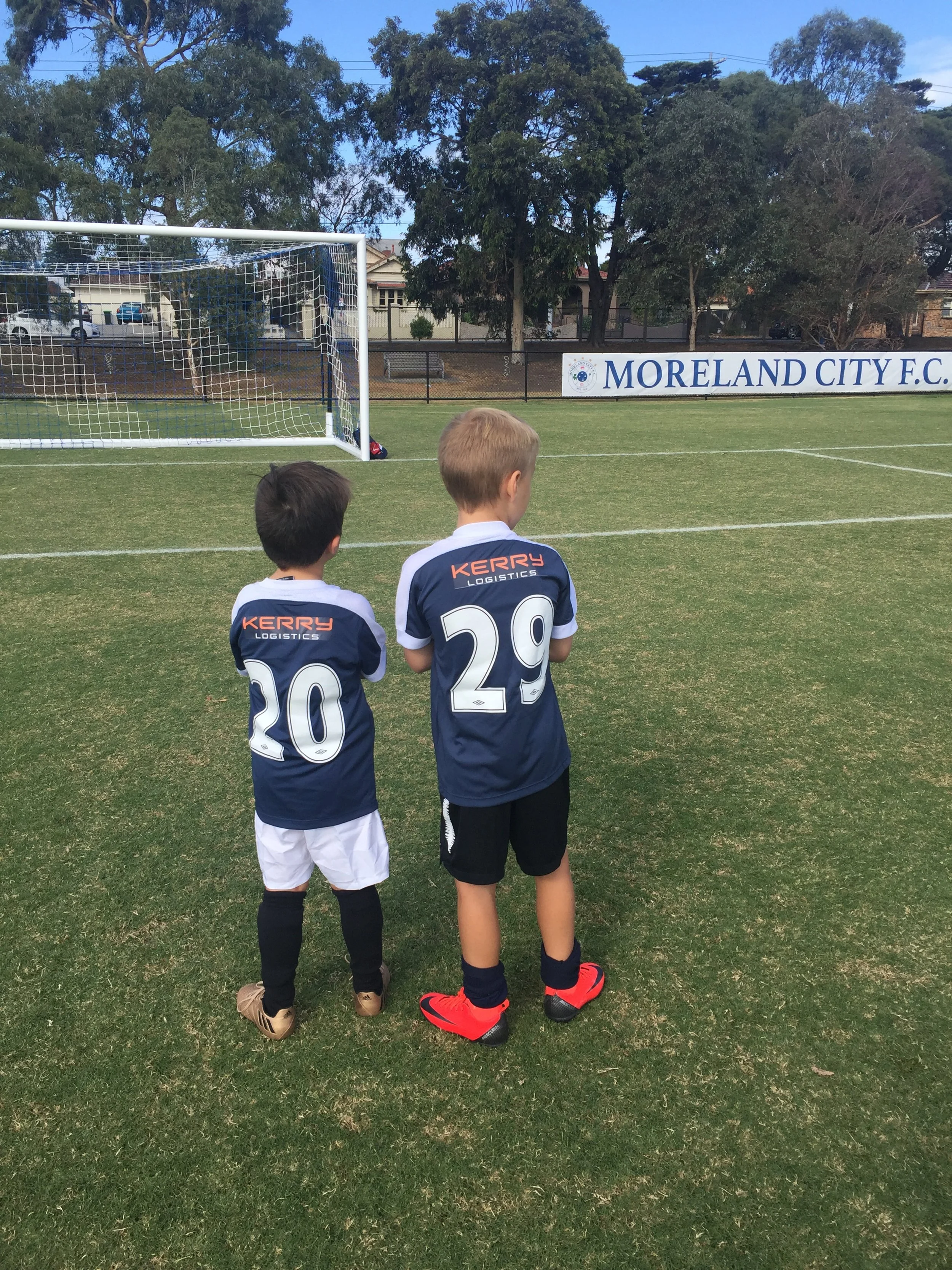 Two young boys in soccer uniforms standing on a soccer field, facing a goal, with trees and a sign reading 'Moreland City F.C.' in the background.