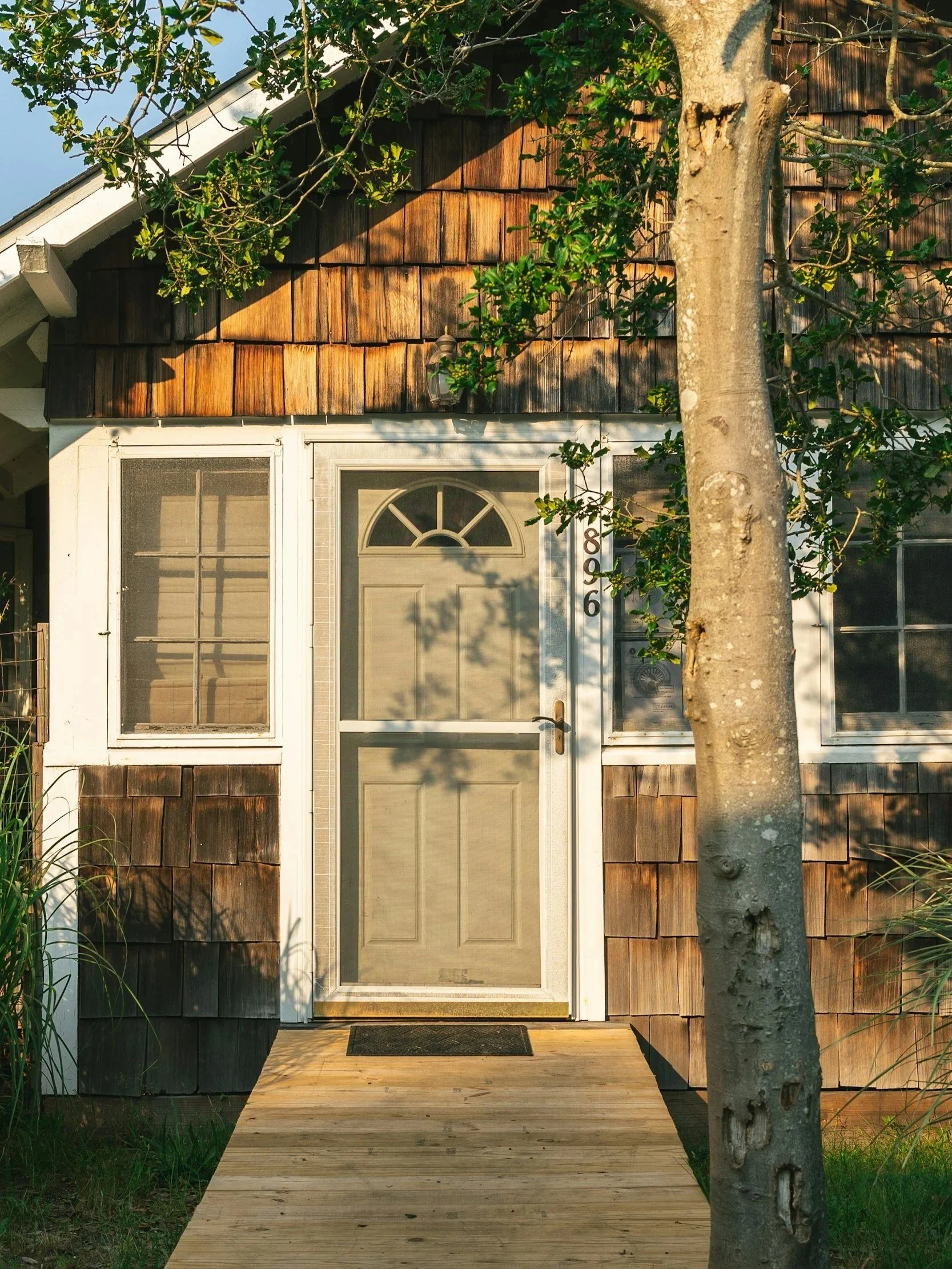 Front view of a house with a wooden walkway leading to a screened door, surrounded by windows and a tree in front.