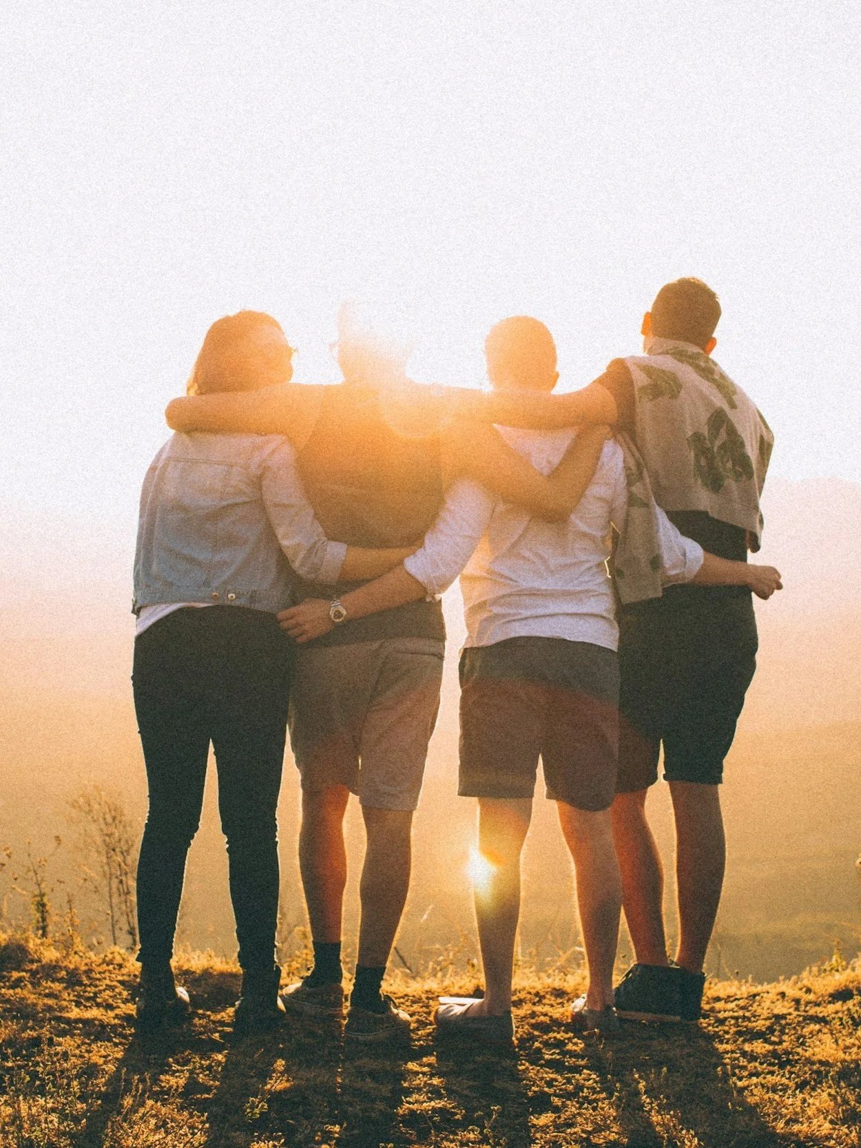 Group of five friends standing together with arms around each other during sunset, viewed from the back.