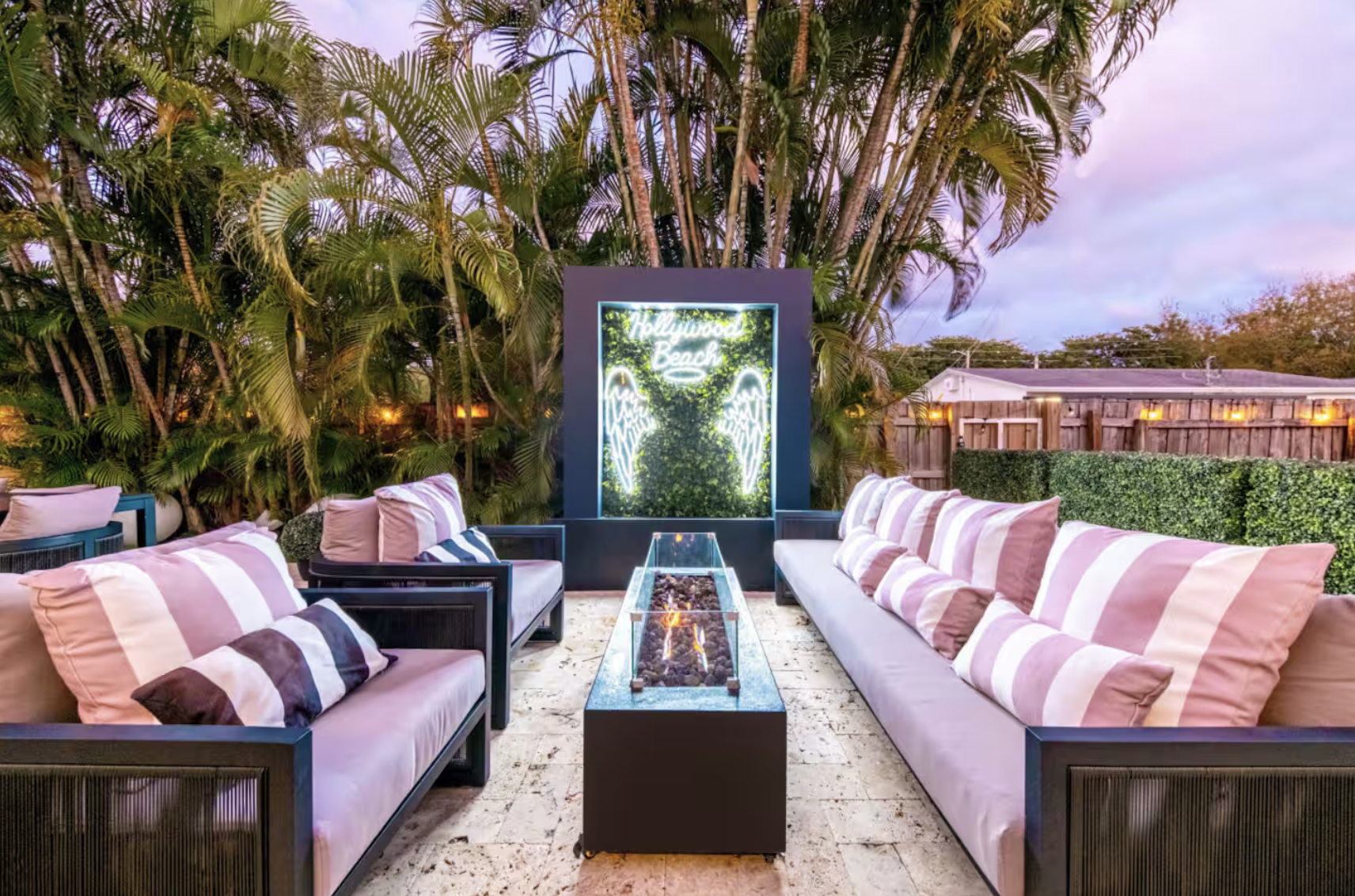 Outdoor patio with two sofas and cushions facing each other, a fire pit table in the center, lush green plants and trees background, neon sign that reads 'Hollywood Beach' with angel wings.
