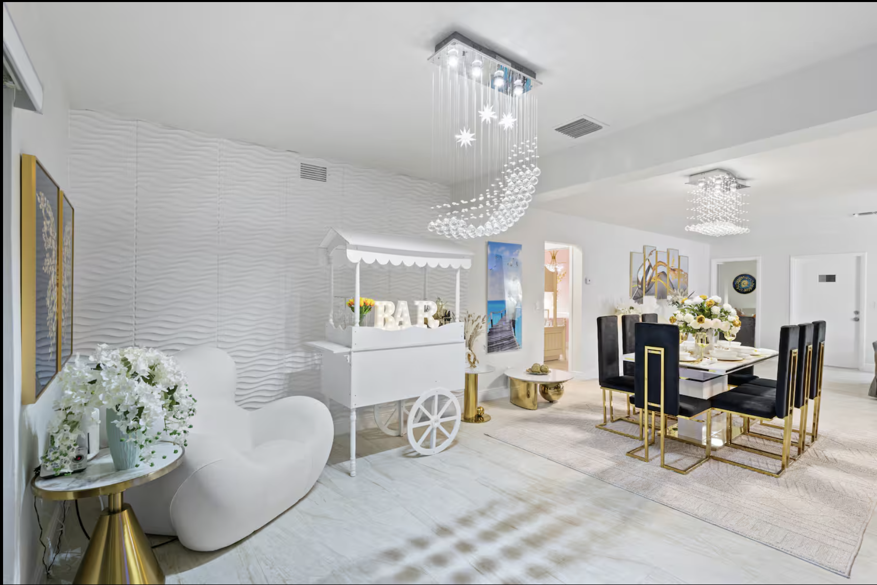 Elegant dining area with black and gold chairs around a white table, floral centerpiece, white textured wall, modern chandeliers, and a white and gold bar cart.