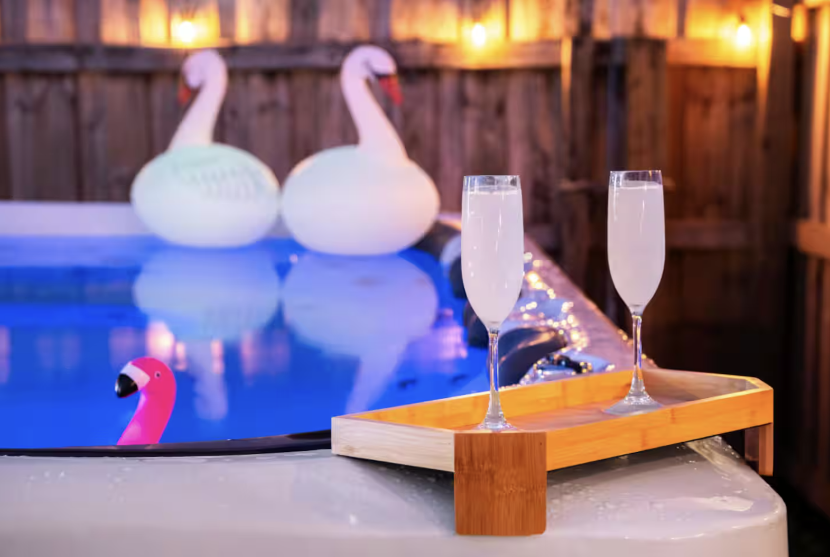 Two swan-shaped pool floats in a swimming pool with a pink flamingo float nearby, illuminated by warm string lights on a wooden fence, with a tray of two champagne glasses on the pool deck.