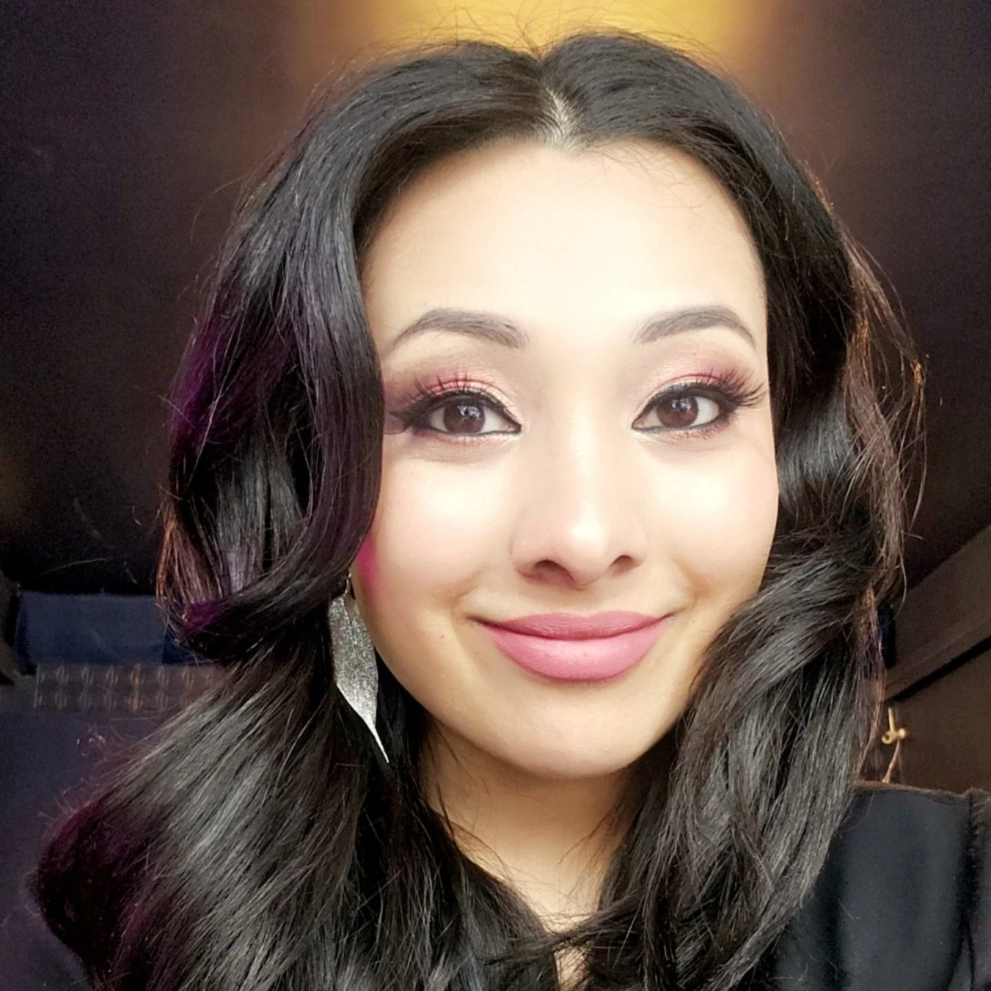 A woman with long, dark wavy hair, wearing makeup including pink eyeshadow and lipstick, and silver earrings, smiling at the camera against a blurred indoor background.