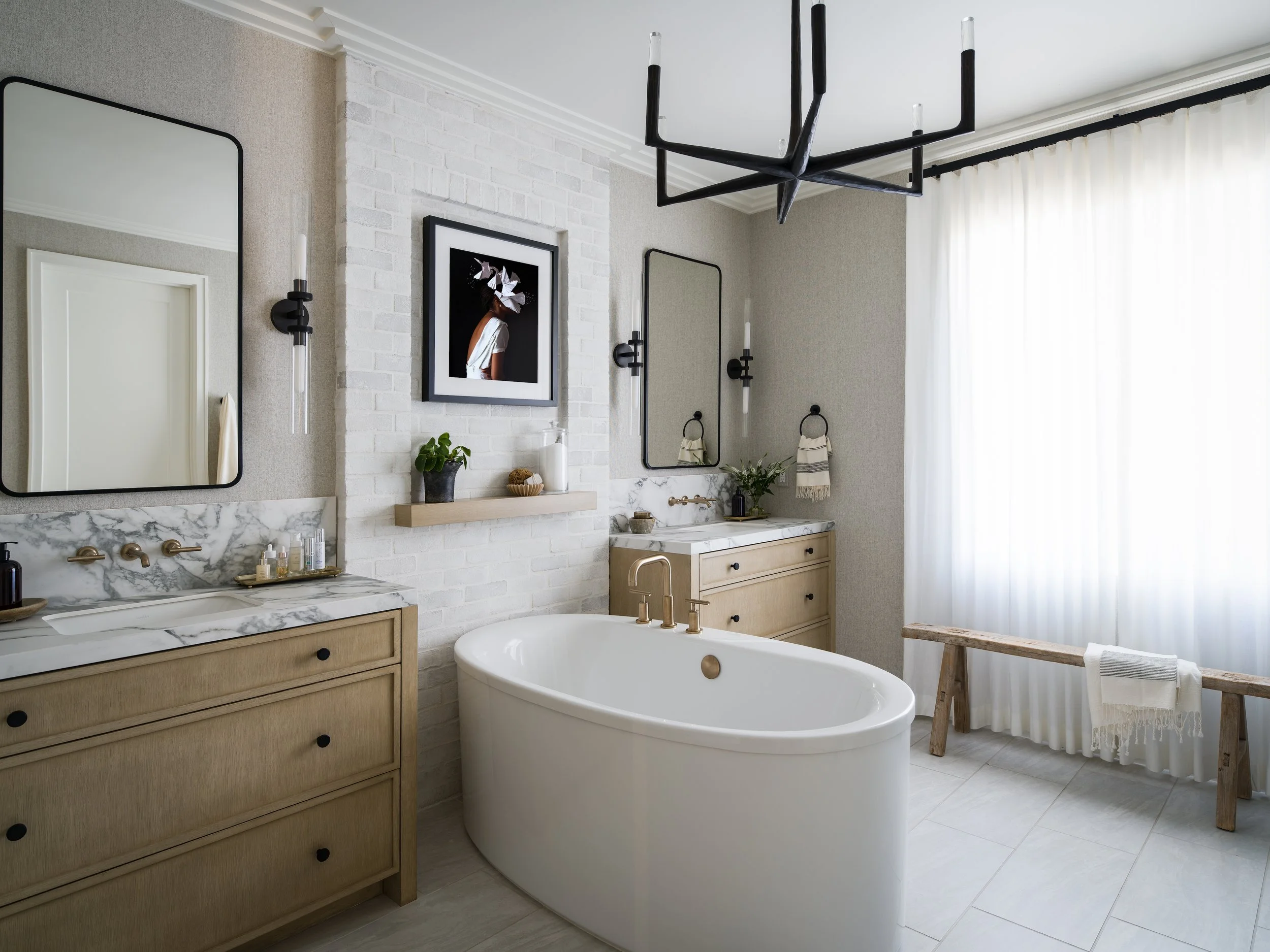 A bathroom with a freestanding bathtub, a wooden vanity with a marble countertop, mirrors, black wall-mounted lights, framed artwork, plants, and a window with white curtains.