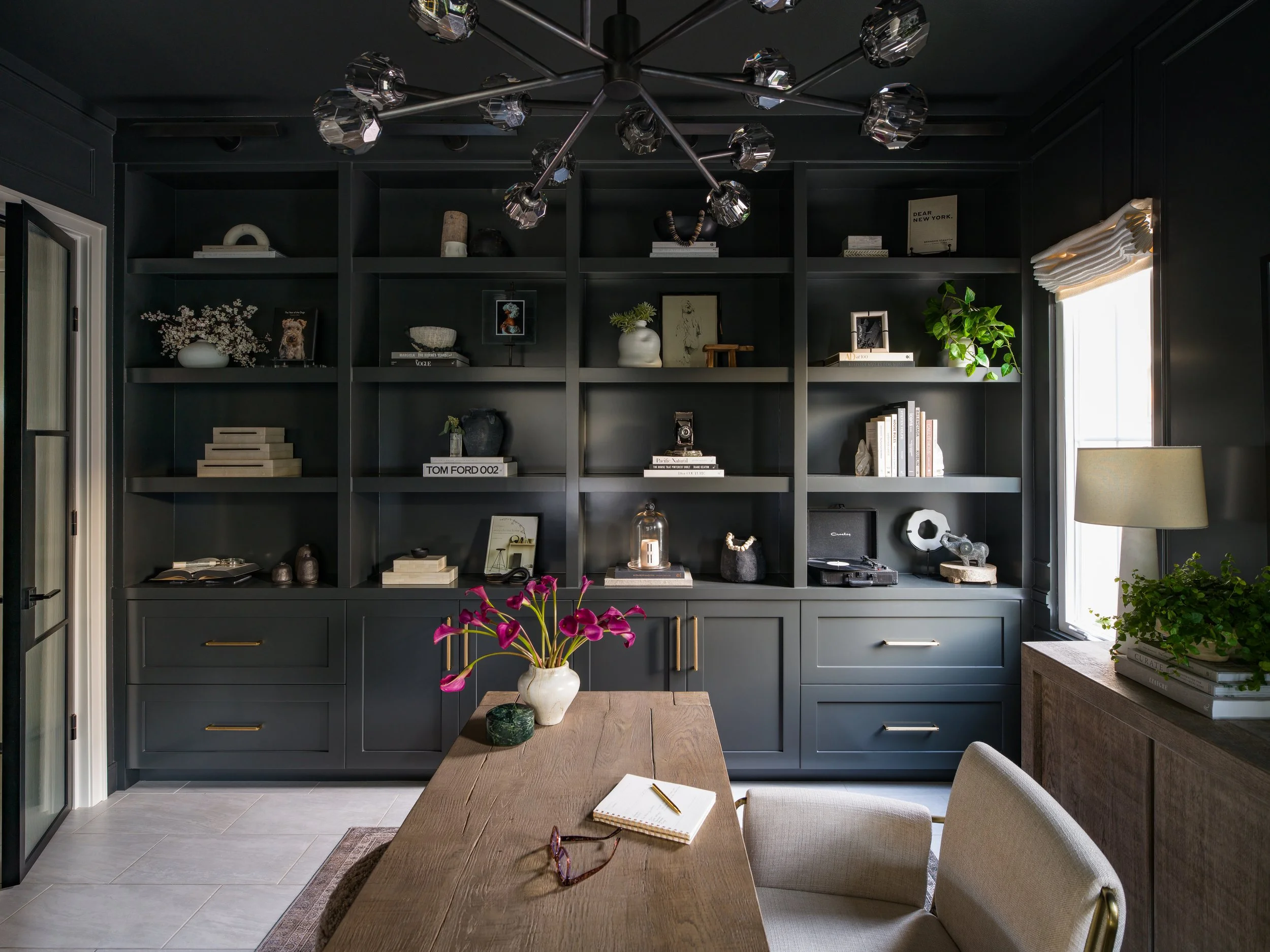 A room with a black bookshelf wall filled with decorative items, books, and plants. A wooden dining table in the foreground has a white vase with pink flowers and an open notebook with glasses. To the right, a window with a white shade lets in natura