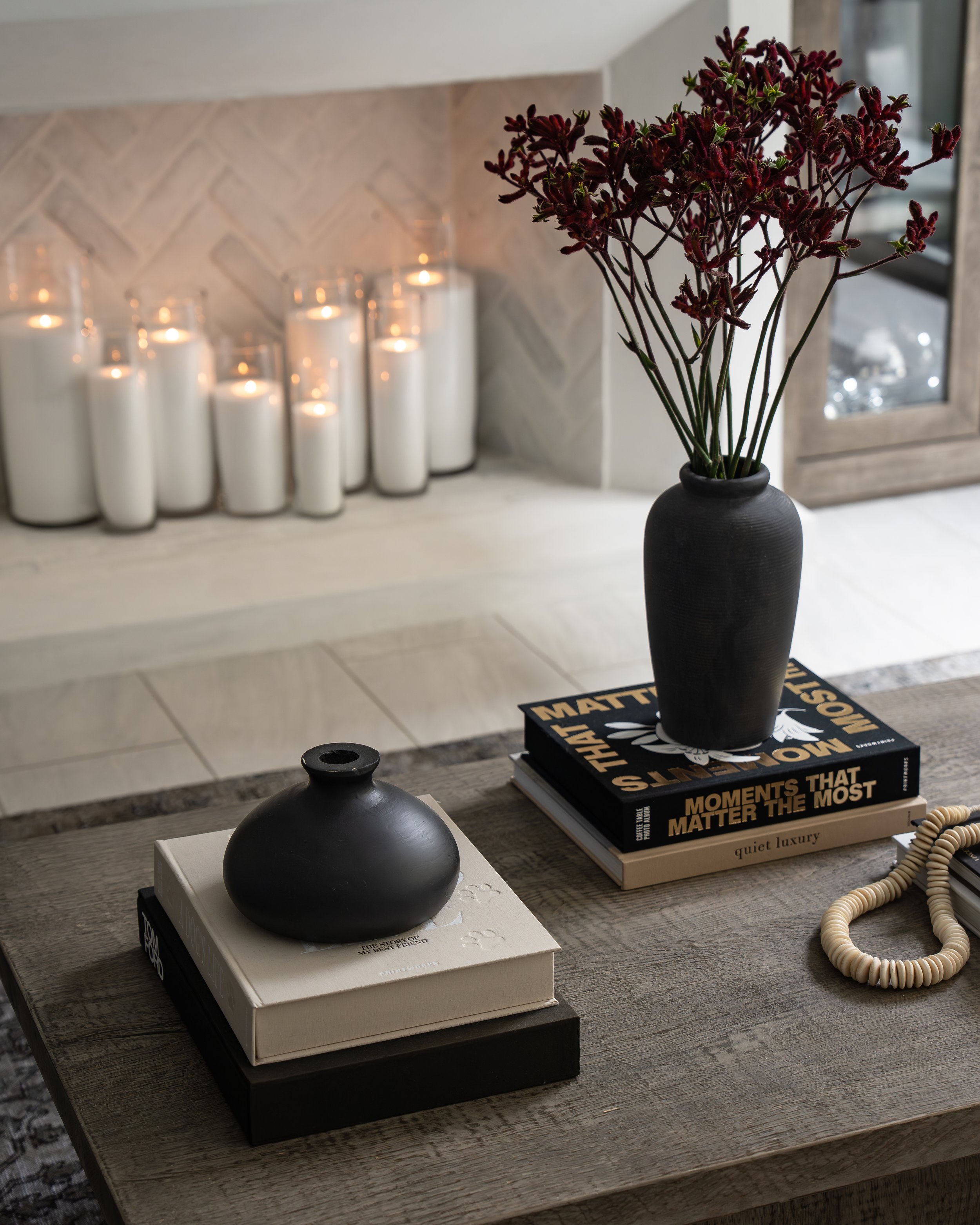 A black vase with dark red flowers on a table, with candles in glass jars and books in the background.