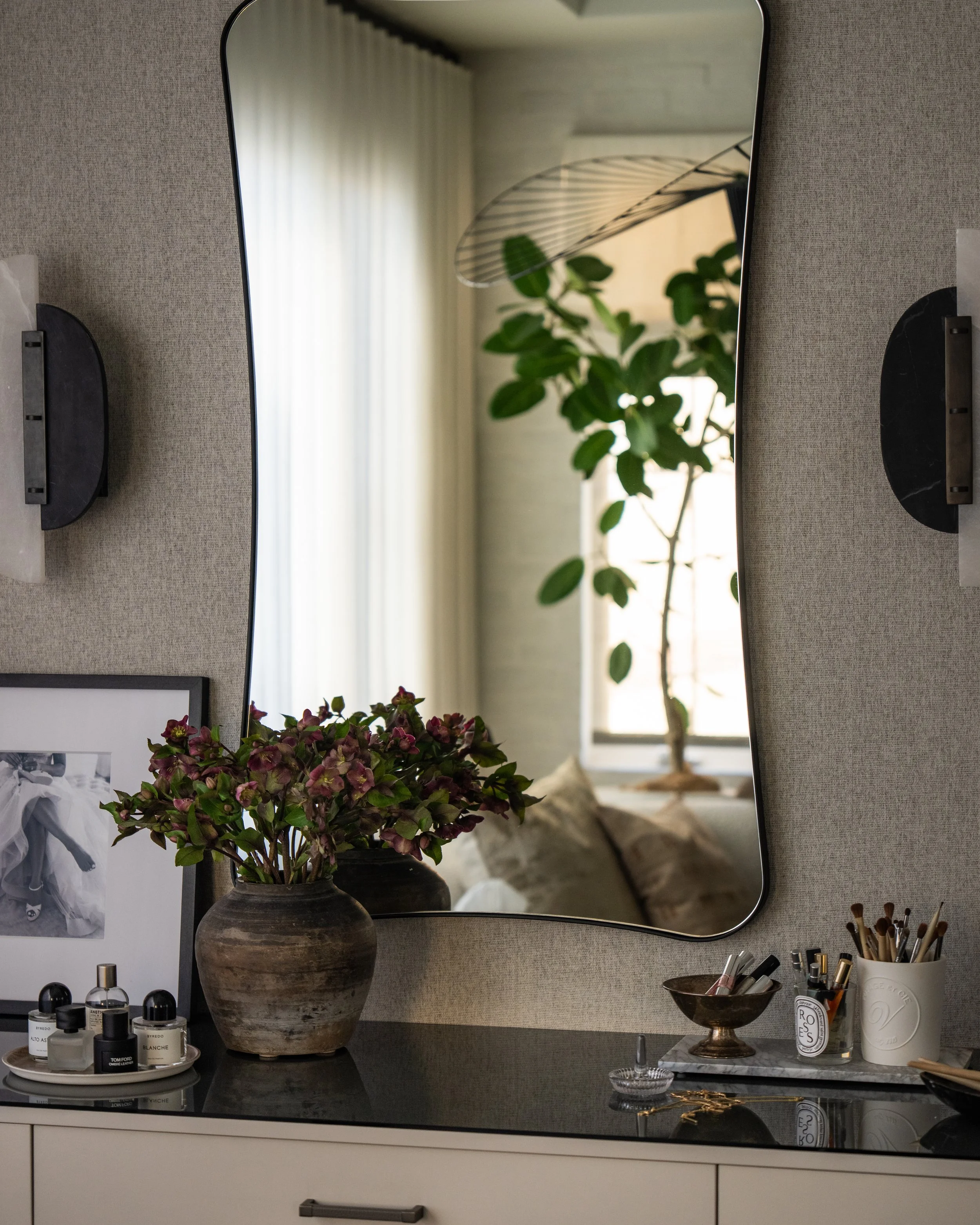 A mirror reflecting green leaves from a plant, with a window and white curtain in the background. The mirror is mounted on a grey wall above a black countertop. On the counter, there are various toiletries, including small bottles, a tray, and a vase