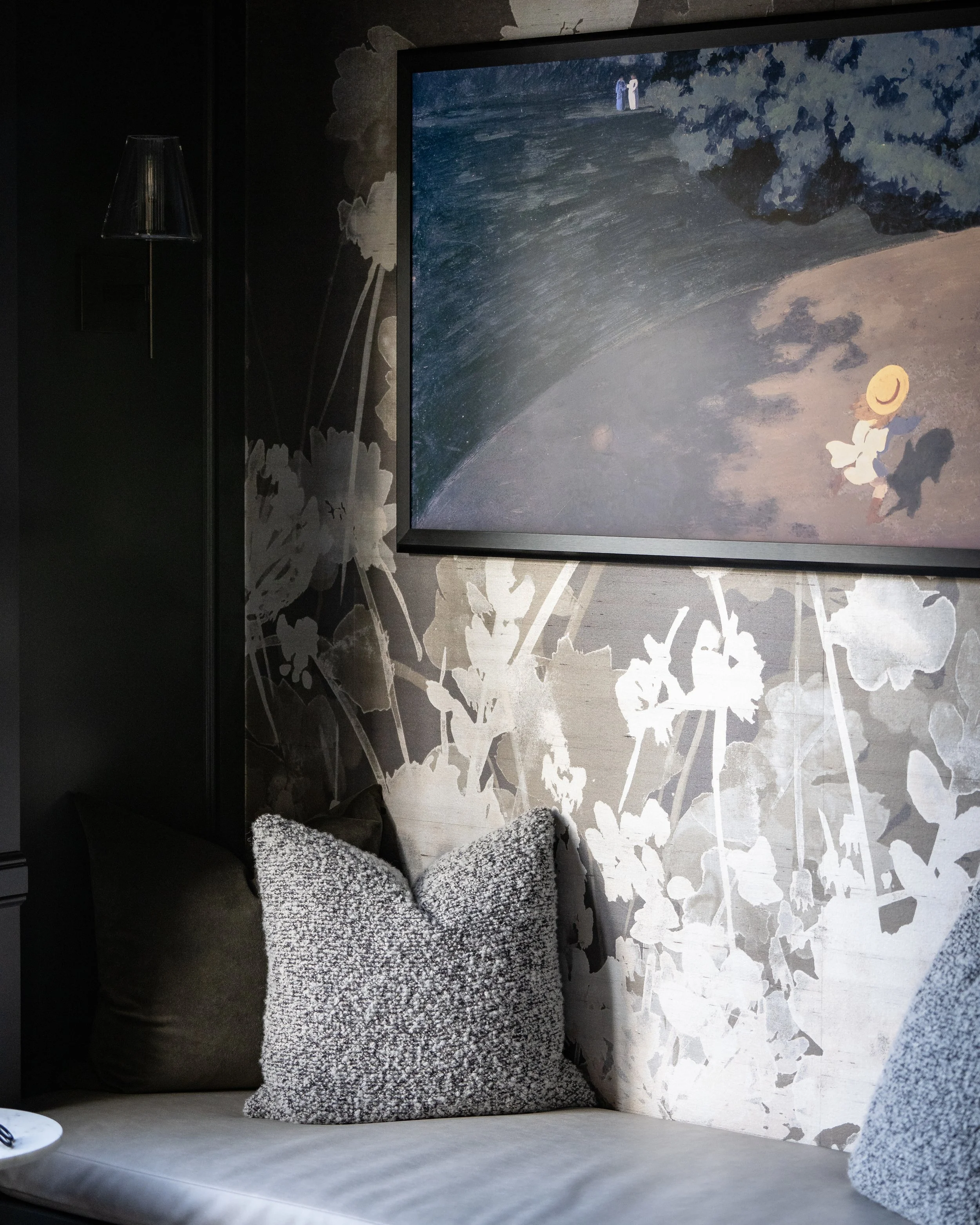 Interior scene of a living room with a grey couch adorned with two patterned throw pillows, a large framed artwork on the wall depicting a beach scene with people and a hat, and a white wall with floral stencil designs.
