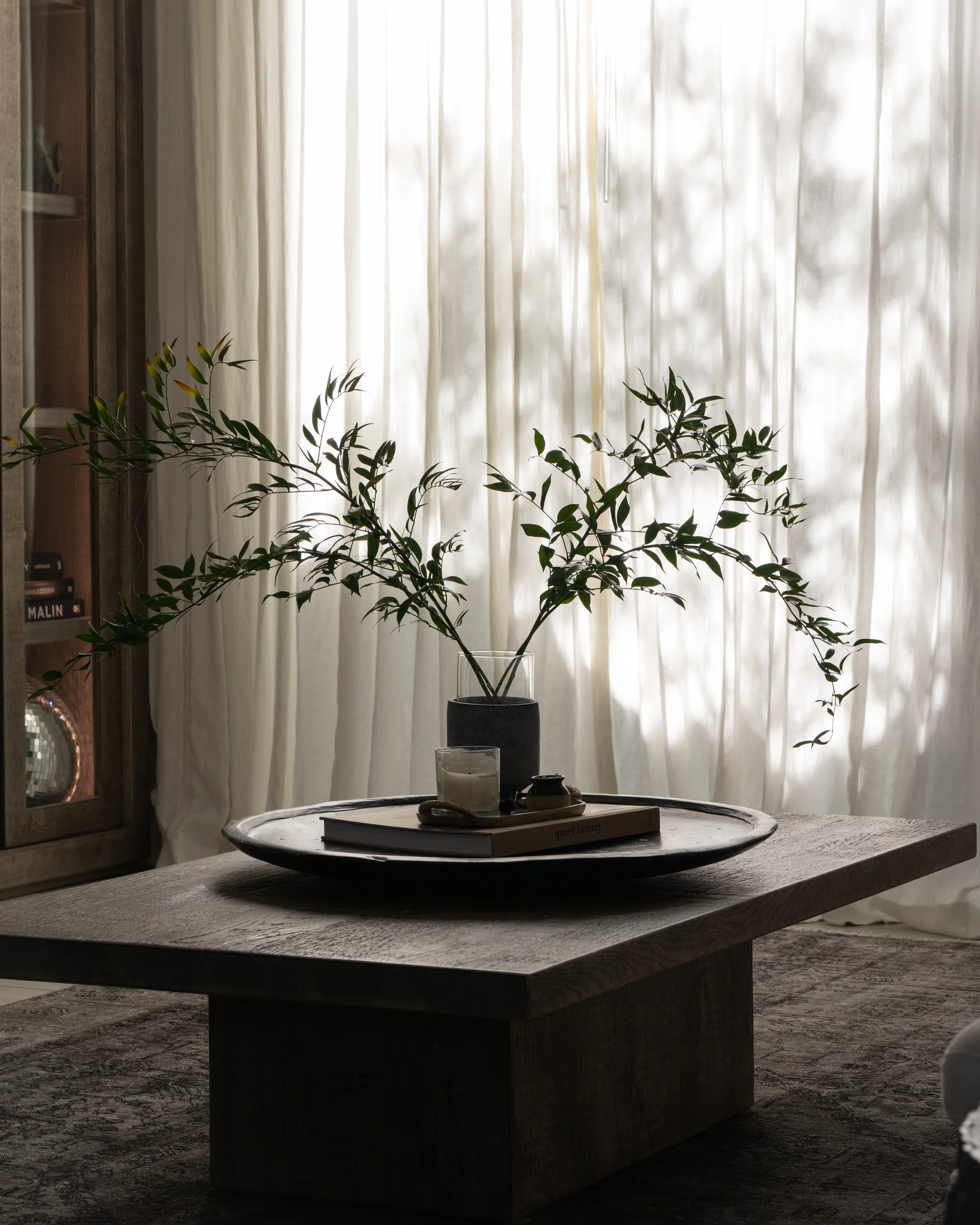 Living room with a wooden table in the center, decorated with a tray holding candles, a small plant, and a book titled 'quiet luxury'. Behind, there's a large window with sheer white curtains allowing natural light to fill the room. To the left, a wo