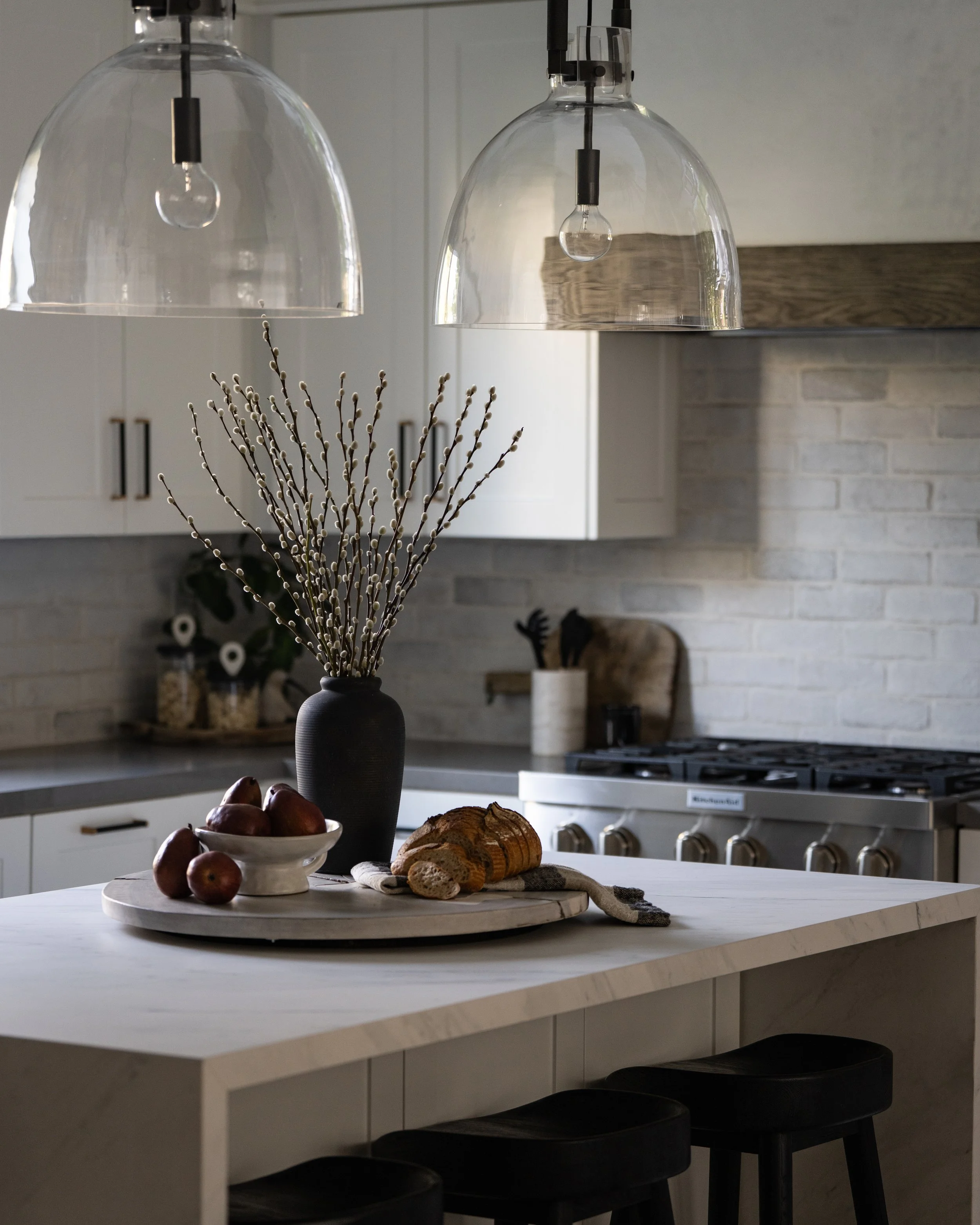 Kitchen interior with two hanging glass pendant lights, a table with a black vase holding pussy willow branches, a bowl of apples, bread, and a dish towel, with a stove and brick wall in the background.