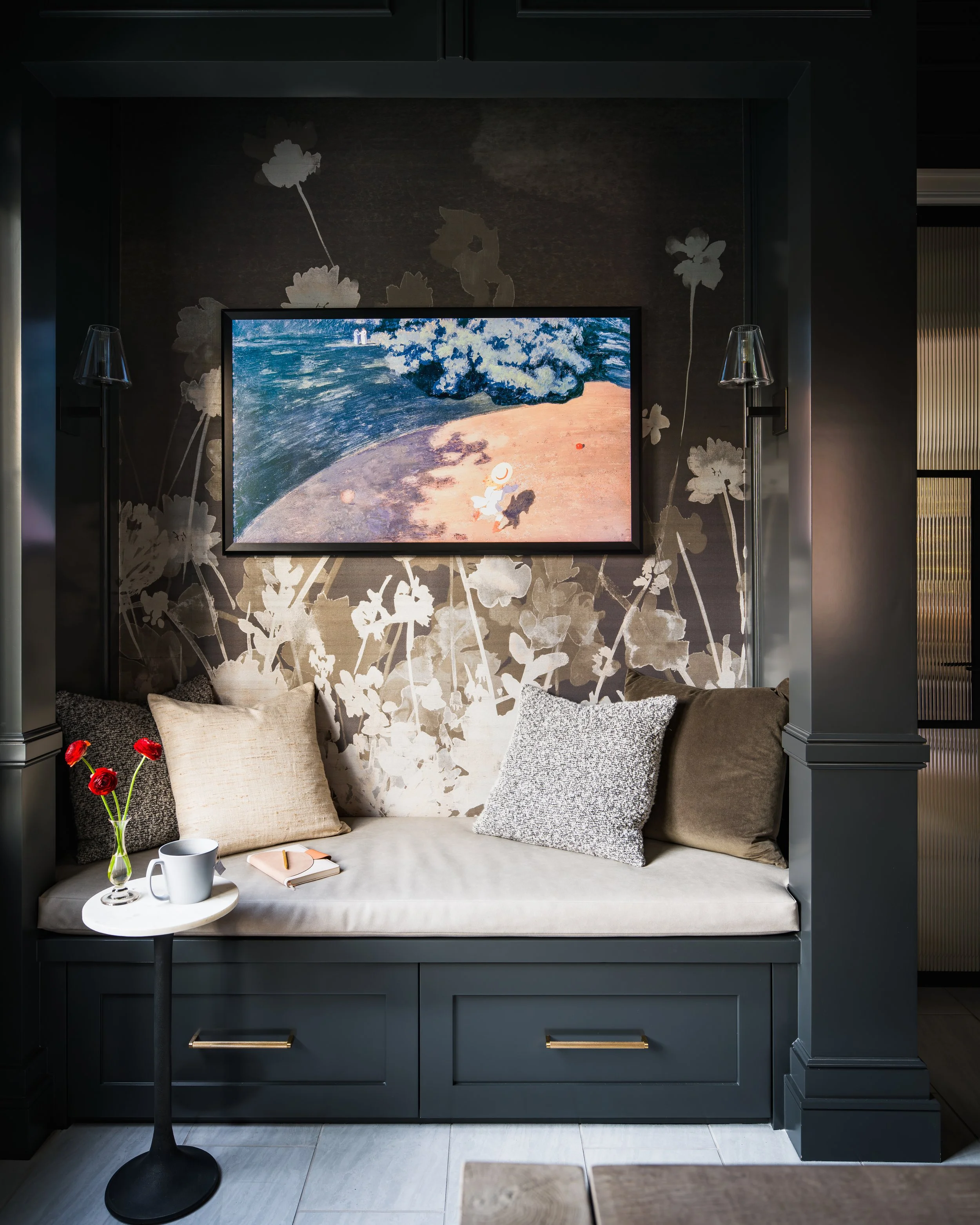 A dark built-in seating nook with floral wallpaper, a framed beach painting, and various pillows. There is a small round side table with a vase of red flowers, a coffee mug, and a notebook. Two wall lamps flank the painting.