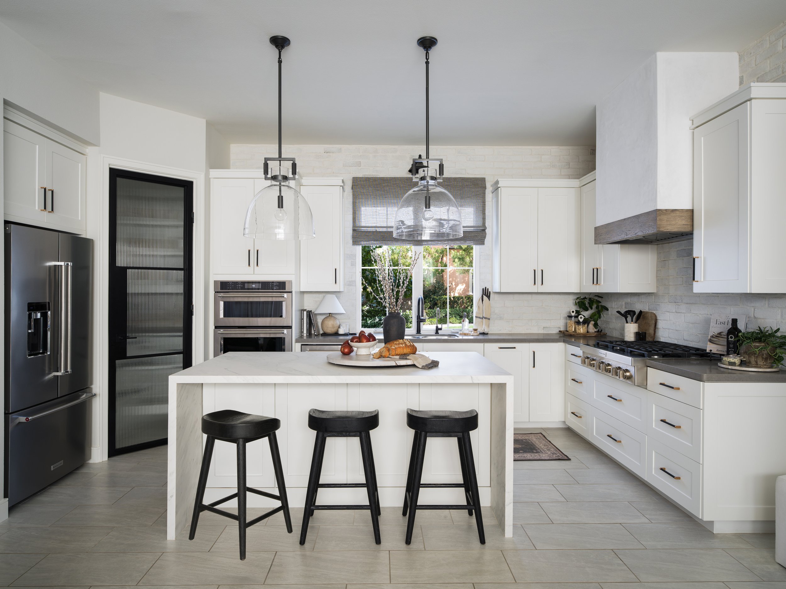 Modern white kitchen with black stools at a island, stainless steel appliances, and a window with greenery outside.