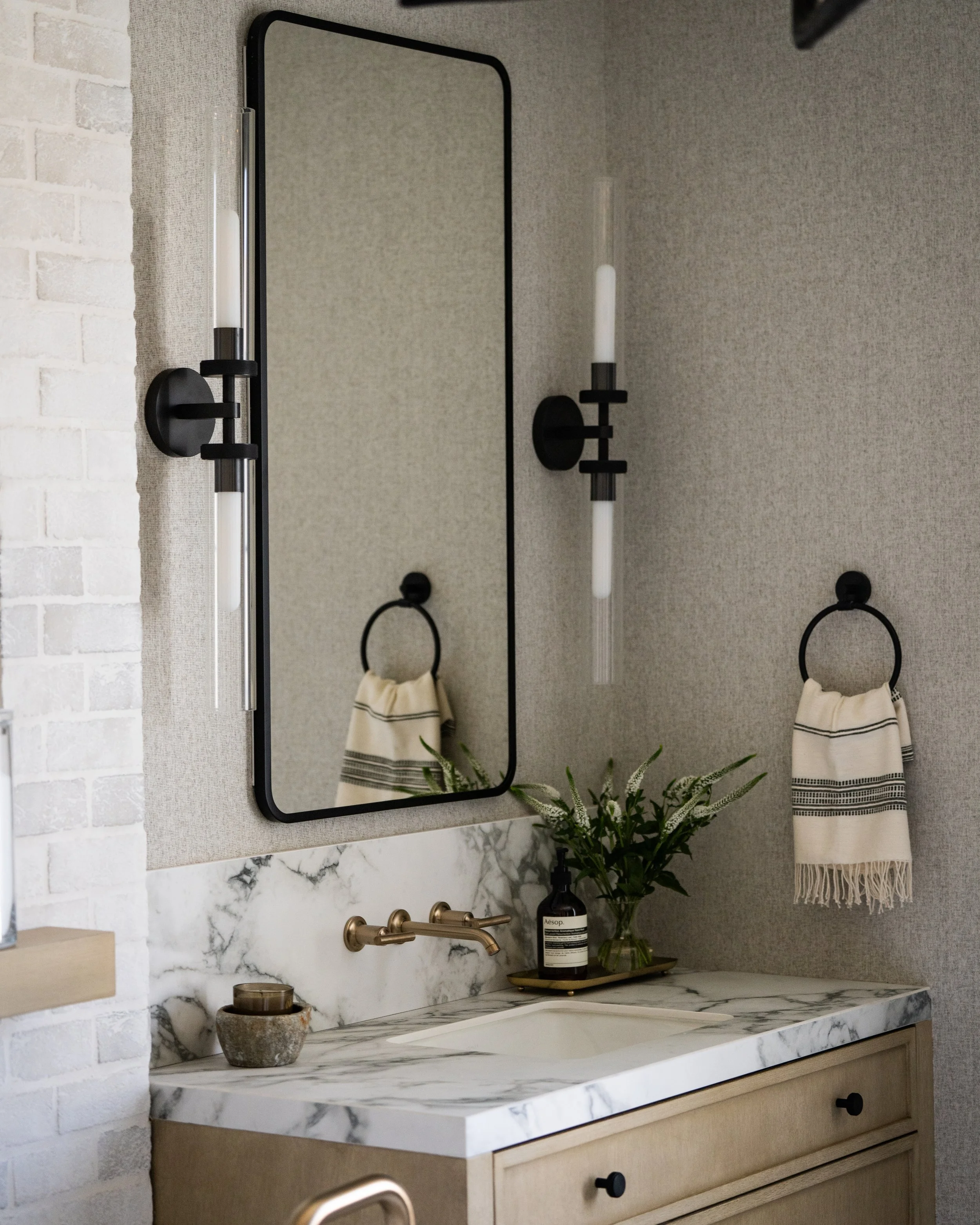 A bathroom sink with a beige wooden vanity, a marble countertop, gold faucet, a small potted plant, a dark bottle of soap, a round marble container, and a rectangular mirror with a black frame. There are two black towel rings with beige and black str