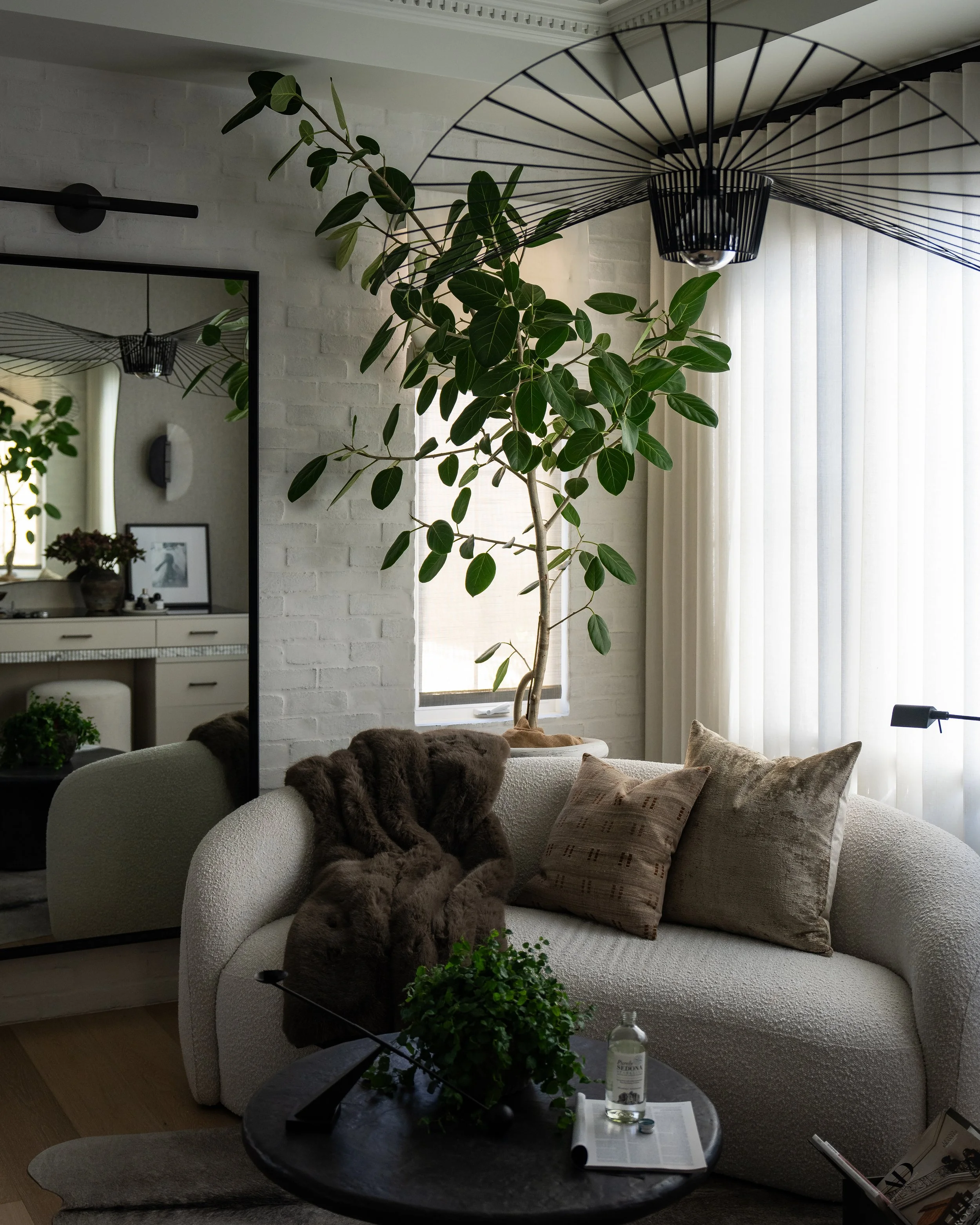 Interior of a living room with a white sofa, grey and beige pillows, a black coffee table with green plants, a water bottle, and some magazines. There is a large window with sheer white curtains, a tall leafy plant, a mirror, a wall light, and a mode