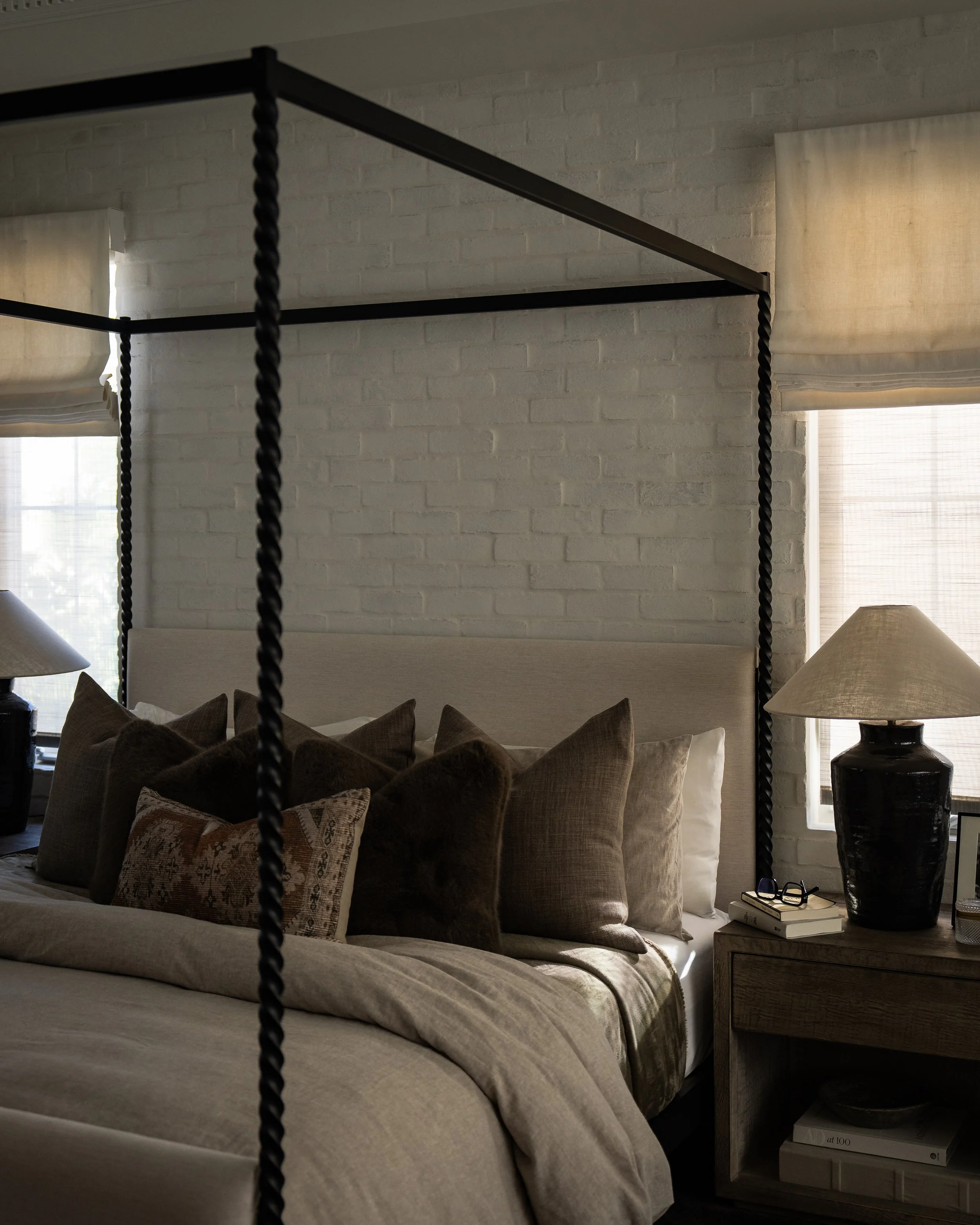 A bedroom with a bed, multiple pillows, two lamps, a nightstand with books, and windows with beige curtains, during daylight.