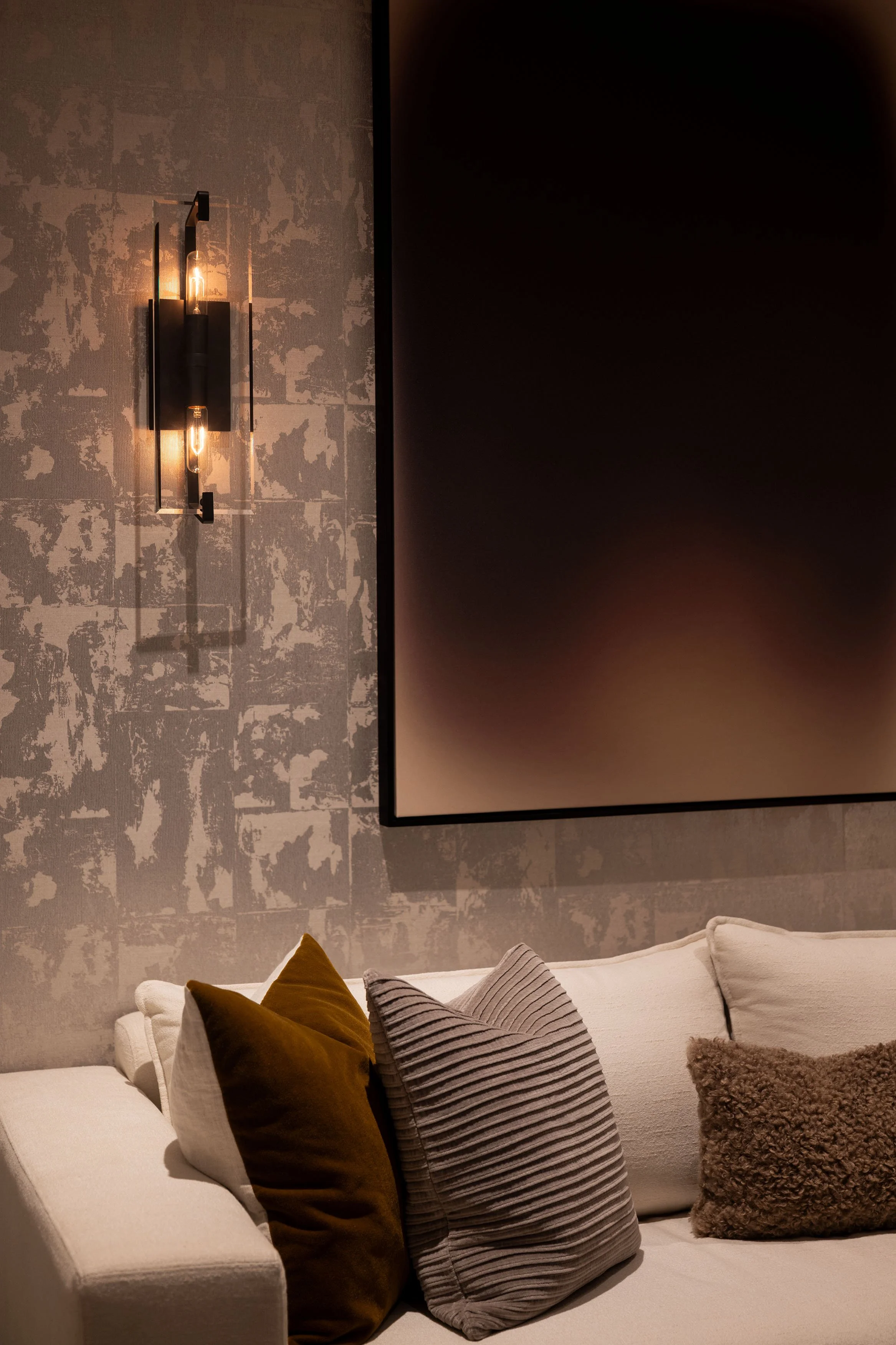 Modern living room with a textured wall, a large framed flat-screen television, a white sofa with various pillows in shades of brown, gray, and beige, and a wall-mounted light fixture with two exposed filament bulbs.