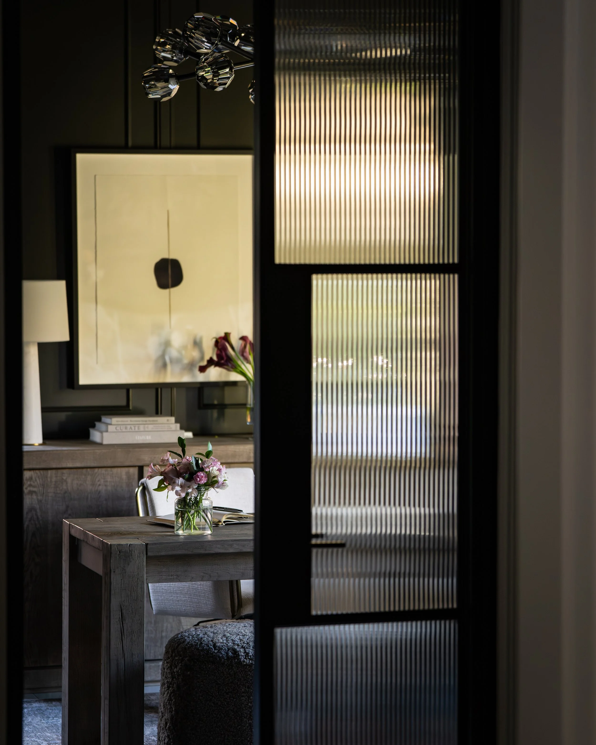 View of a cozy living room seen through a sliding door with textured glass panels. Inside, there is a rustic wooden table with a glass vase holding pink and white flowers, next to a bentwood chair. In the background, a light-colored wall features a l