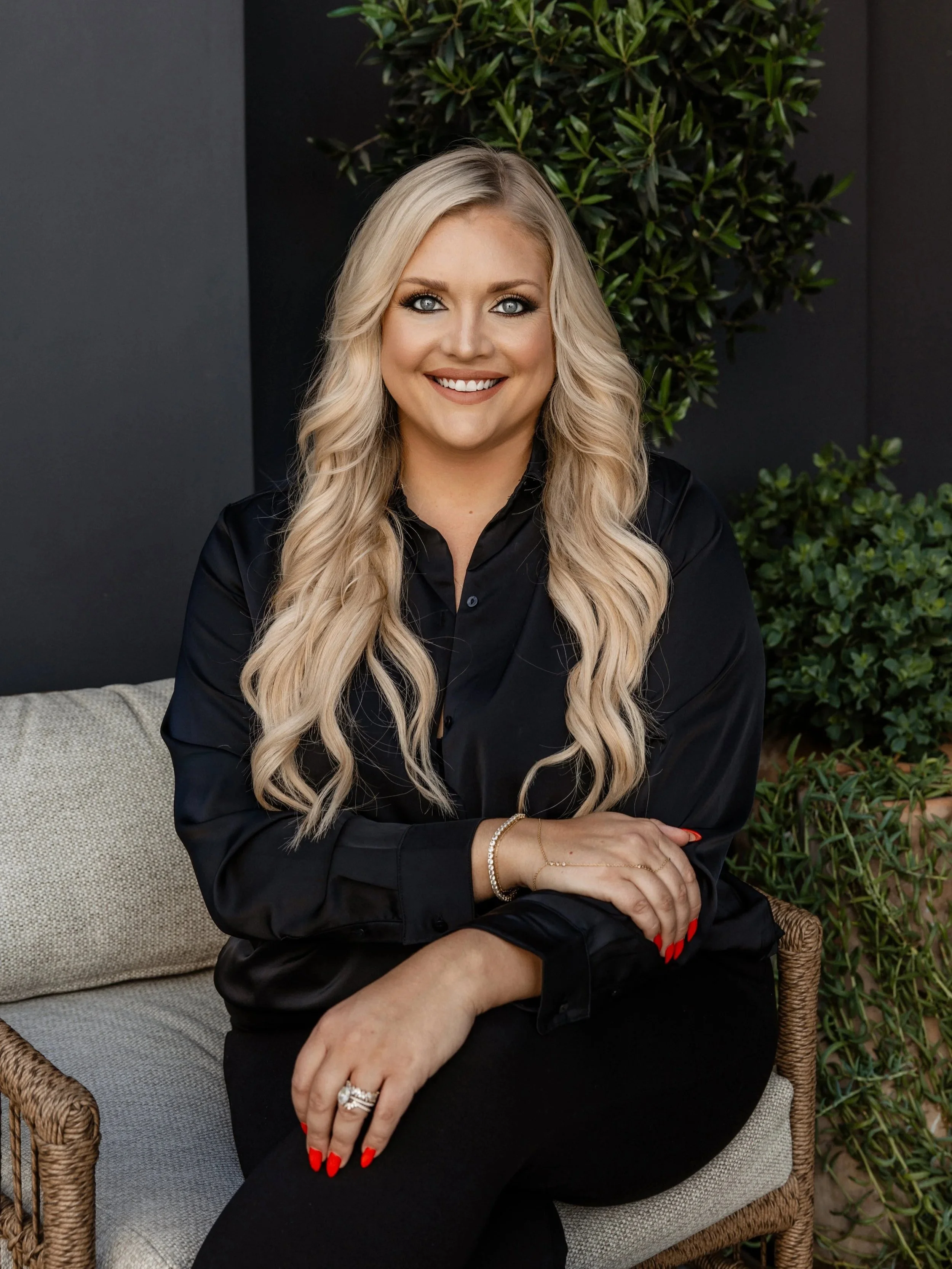 A smiling woman with long blonde wavy hair, wearing a black shirt and red nail polish, sitting on an outdoor wicker chair with a light-colored cushion, surrounded by lush green plants.