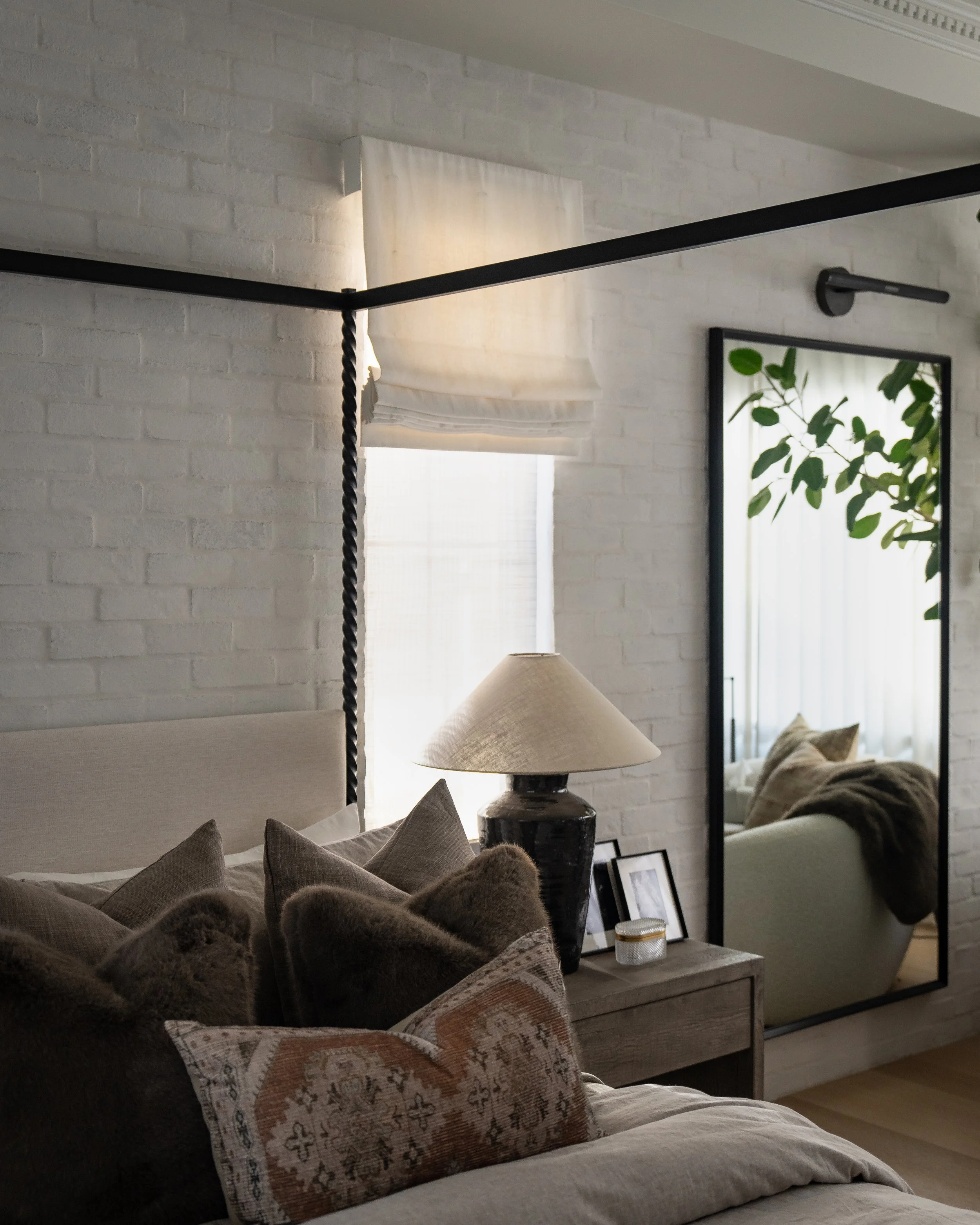 A cozy living room scene with beige sofas, decorative pillows, a side table with a lamp, a large mirror, and a wall with white brick texture. The room has natural light coming through a window with white curtains.