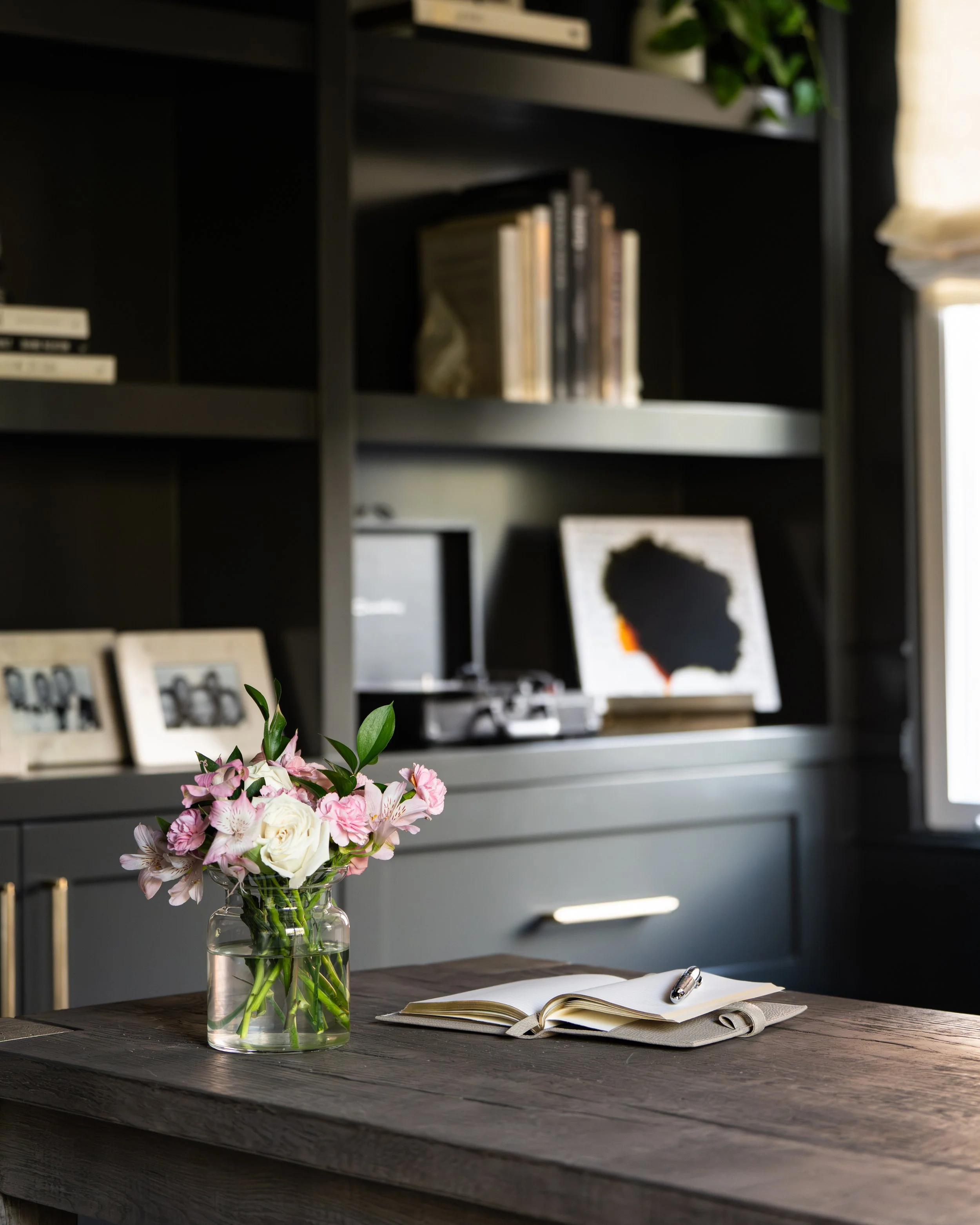 A glass vase filled with pink and white flowers on a dark wooden table, with an open notebook and a silver pen, in front of a gray bookshelf filled with books and a framed picture, in a well-lit room.