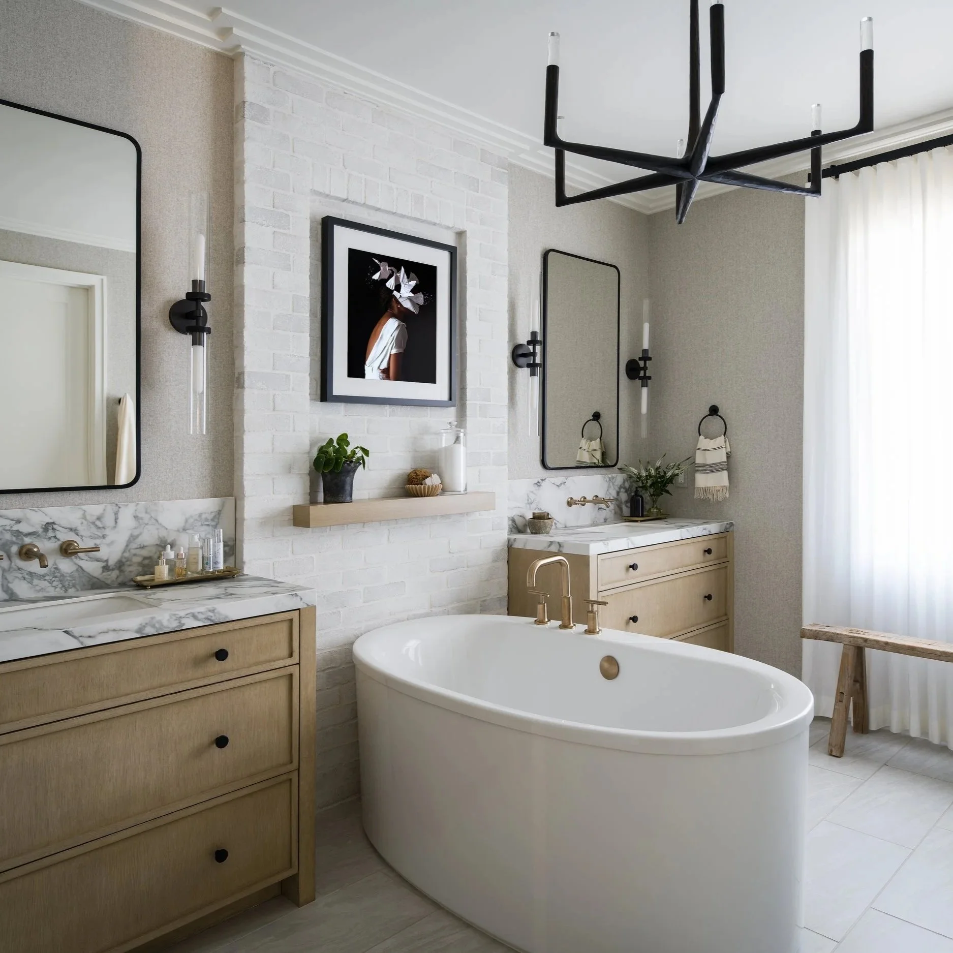 Modern bathroom with white freestanding bathtub, wooden vanity with marble countertop, framed artwork, large mirror, wall sconces, and a window with white curtains.