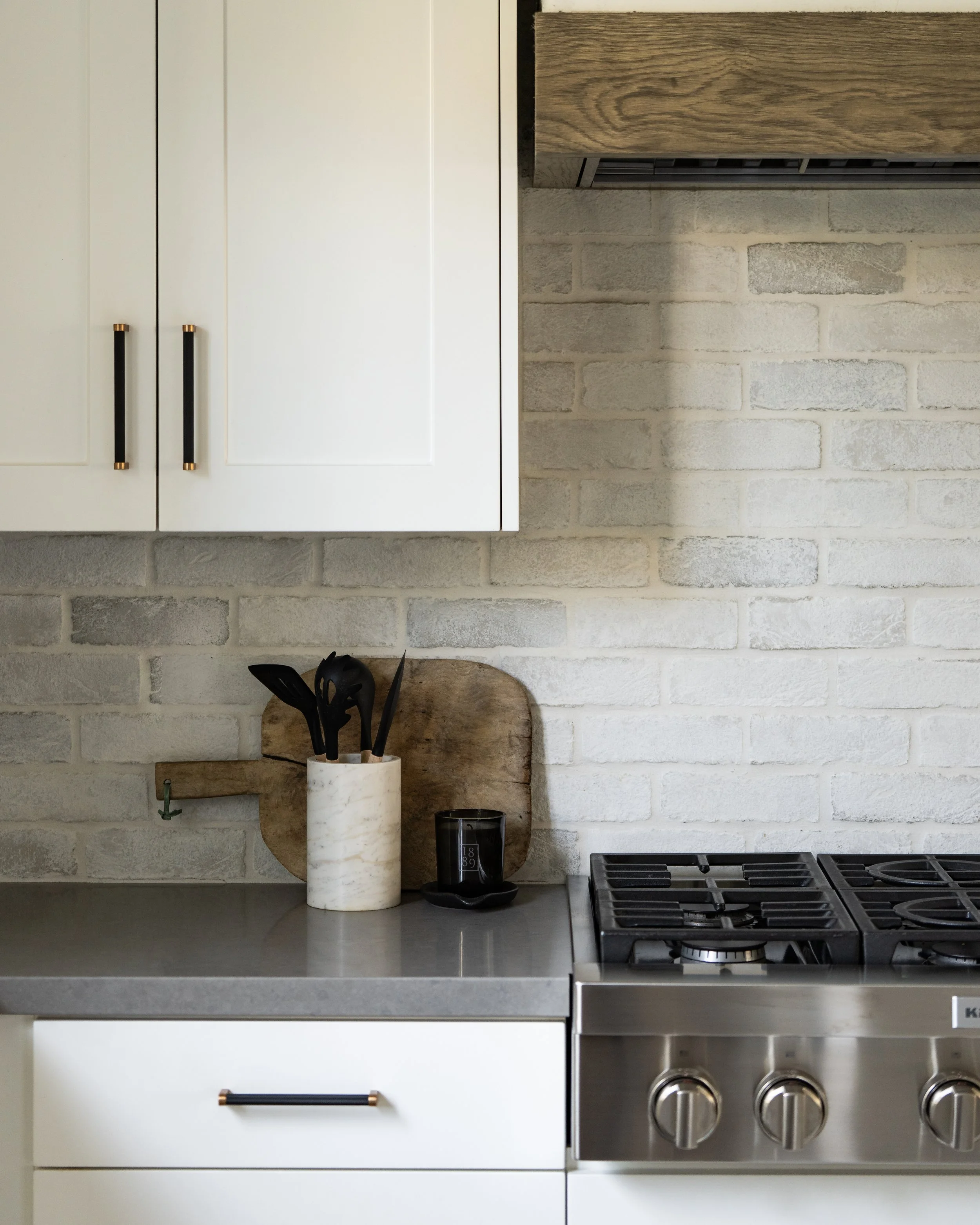 Kitchen countertop with a white cabinet above, a wooden cutting board with black utensils in a white marble container, a black tumbler, and a modern stainless steel gas stove with three knobs in a contemporary kitchen.