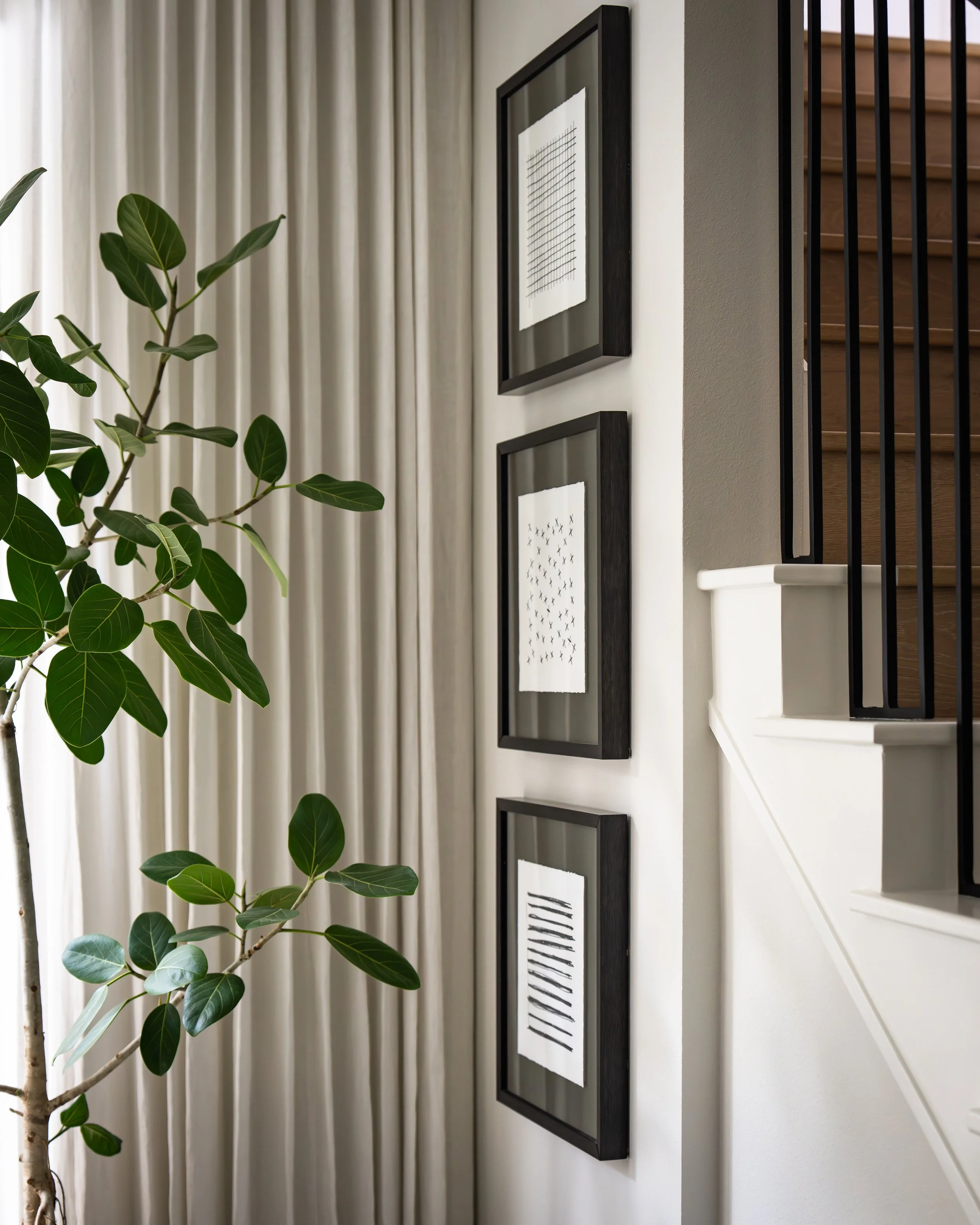 Interior view of a staircase with black metal railings, framed black and white photos on the wall, a plant with broad green leaves, beige curtains on a window, and a light-colored wall.
