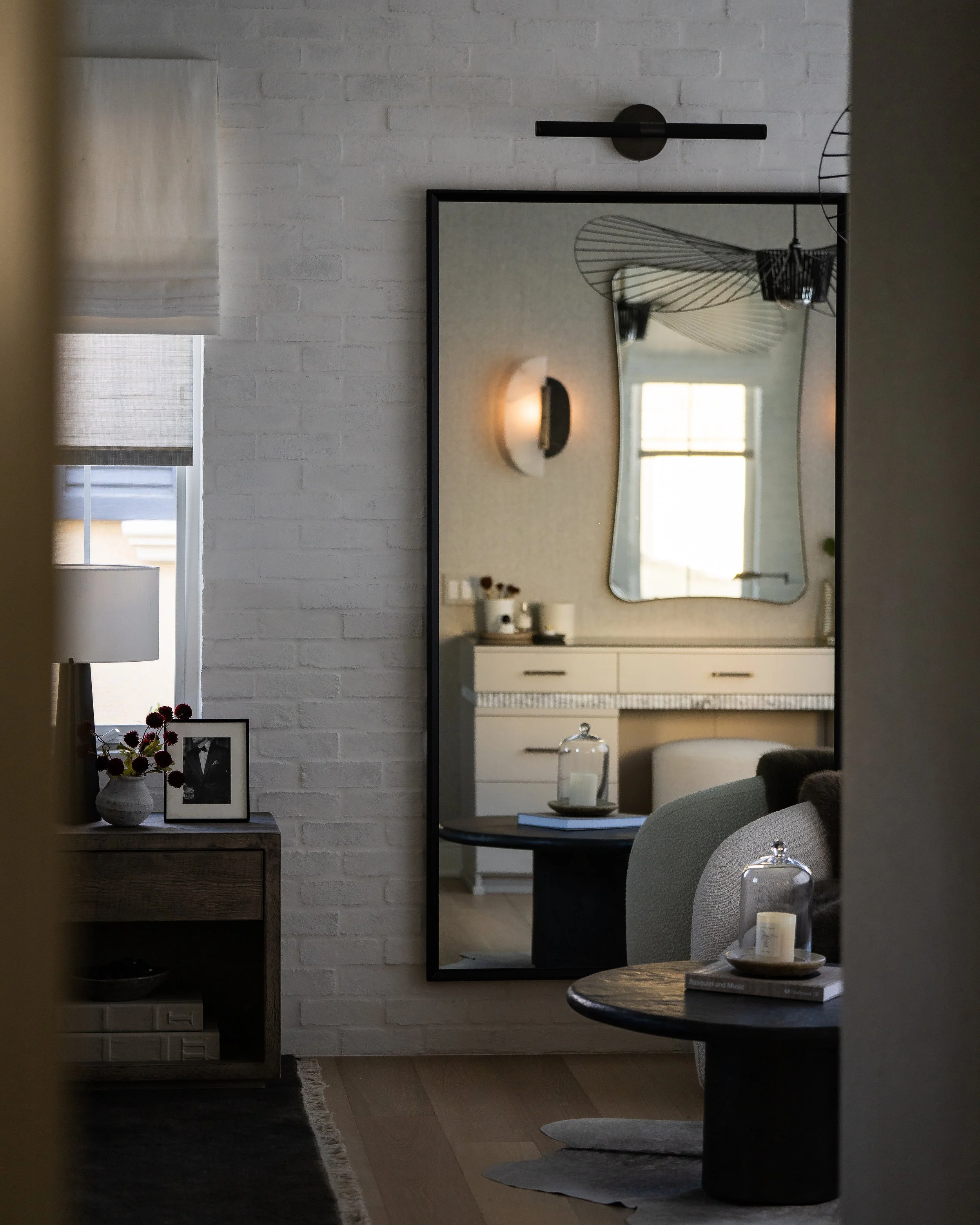 Interior view of a modern room reflecting in a large wall mirror. The room features minimalistic decor including a white brick wall, sleek black and white furniture, a pendant light, window with white blinds, and decorative items like vases, books, a