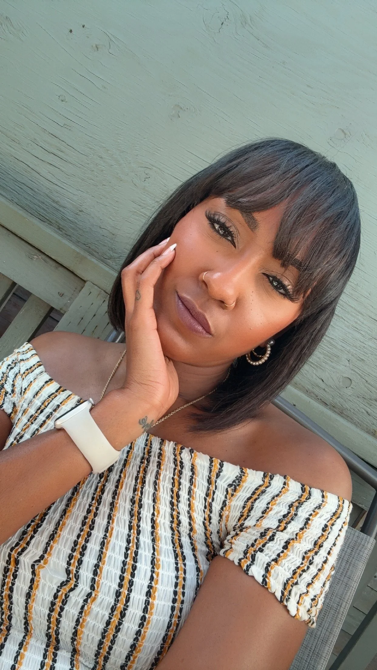 A woman with shoulder-length dark hair and bangs, wearing hoop earrings, a nose ring, and a striped off-the-shoulder top, sitting outdoors against a wooden wall, with her hand resting on her face.