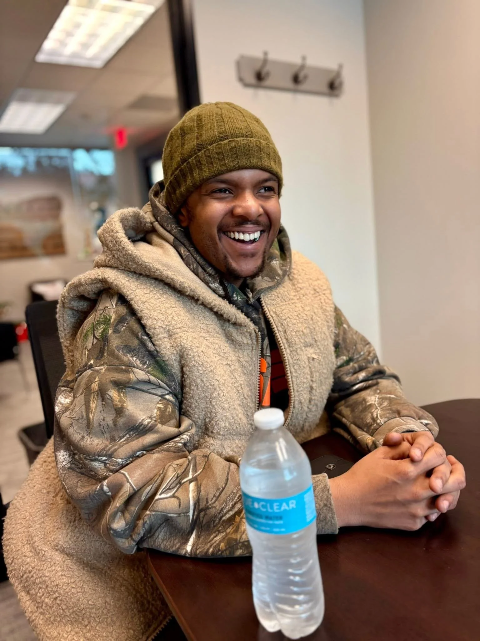 A smiling man wearing a brown beanie and camouflaged jacket sitting at a table with a water bottle and a phone inside a room with ceiling lights and hooks on the wall.