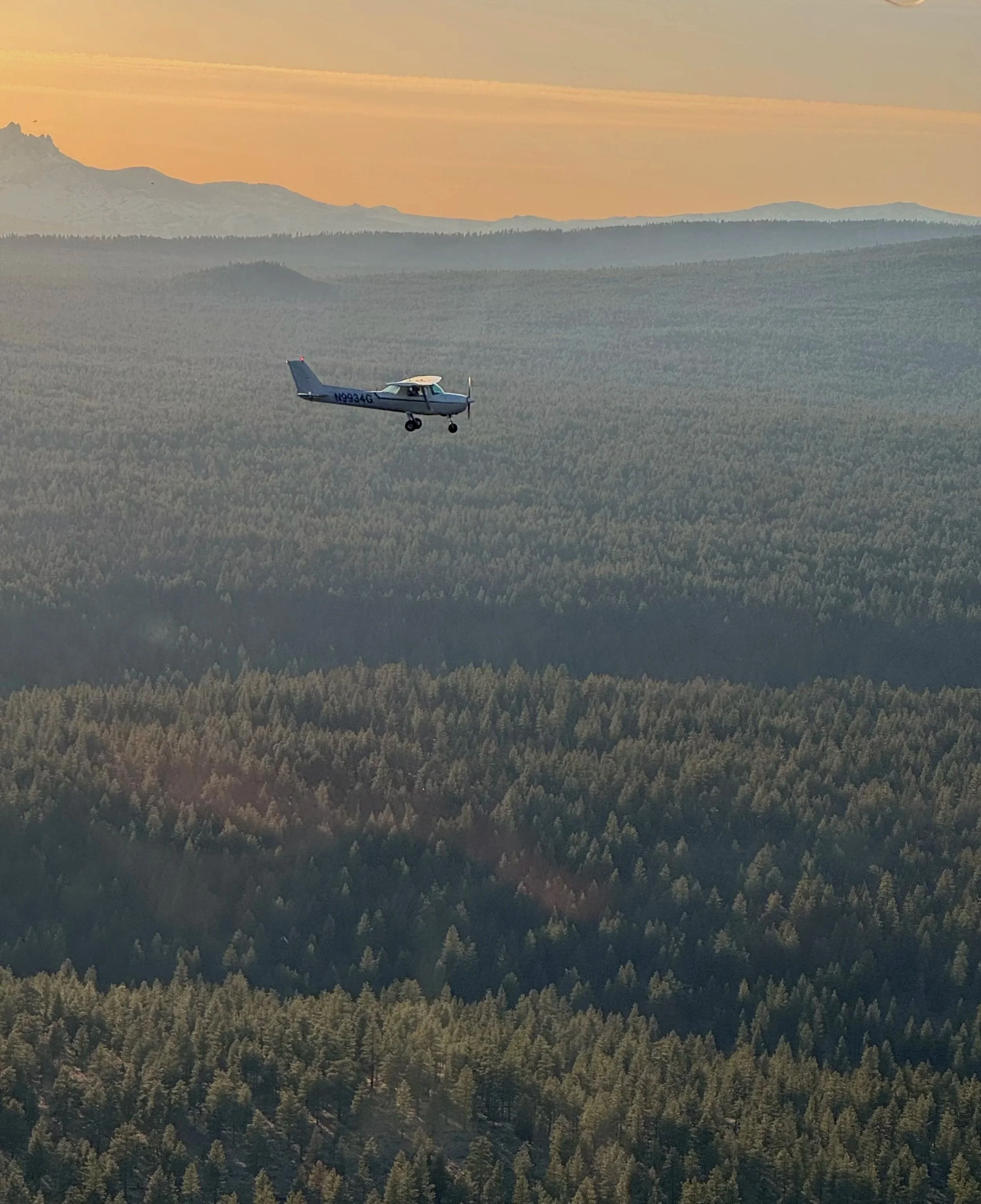 Small airplane flying above a dense forest with mountains in the background during sunset.