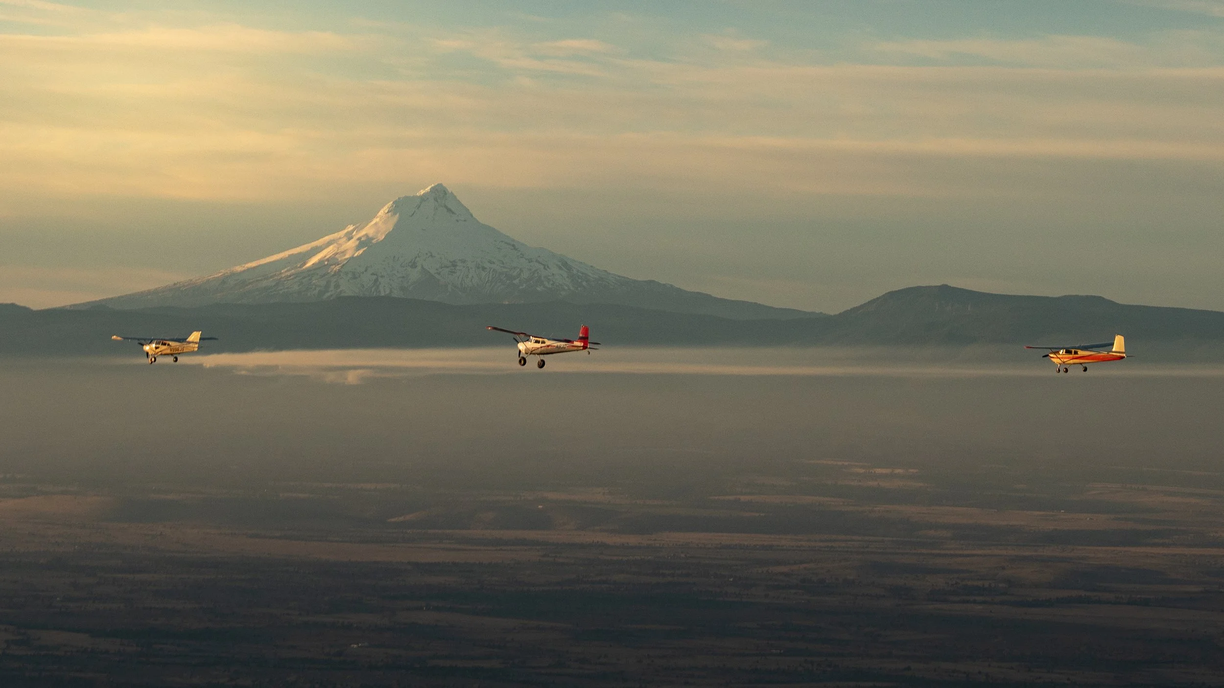 Three small planes flying in formation against a backdrop of Mount St. Helens, with a partly cloudy sky.