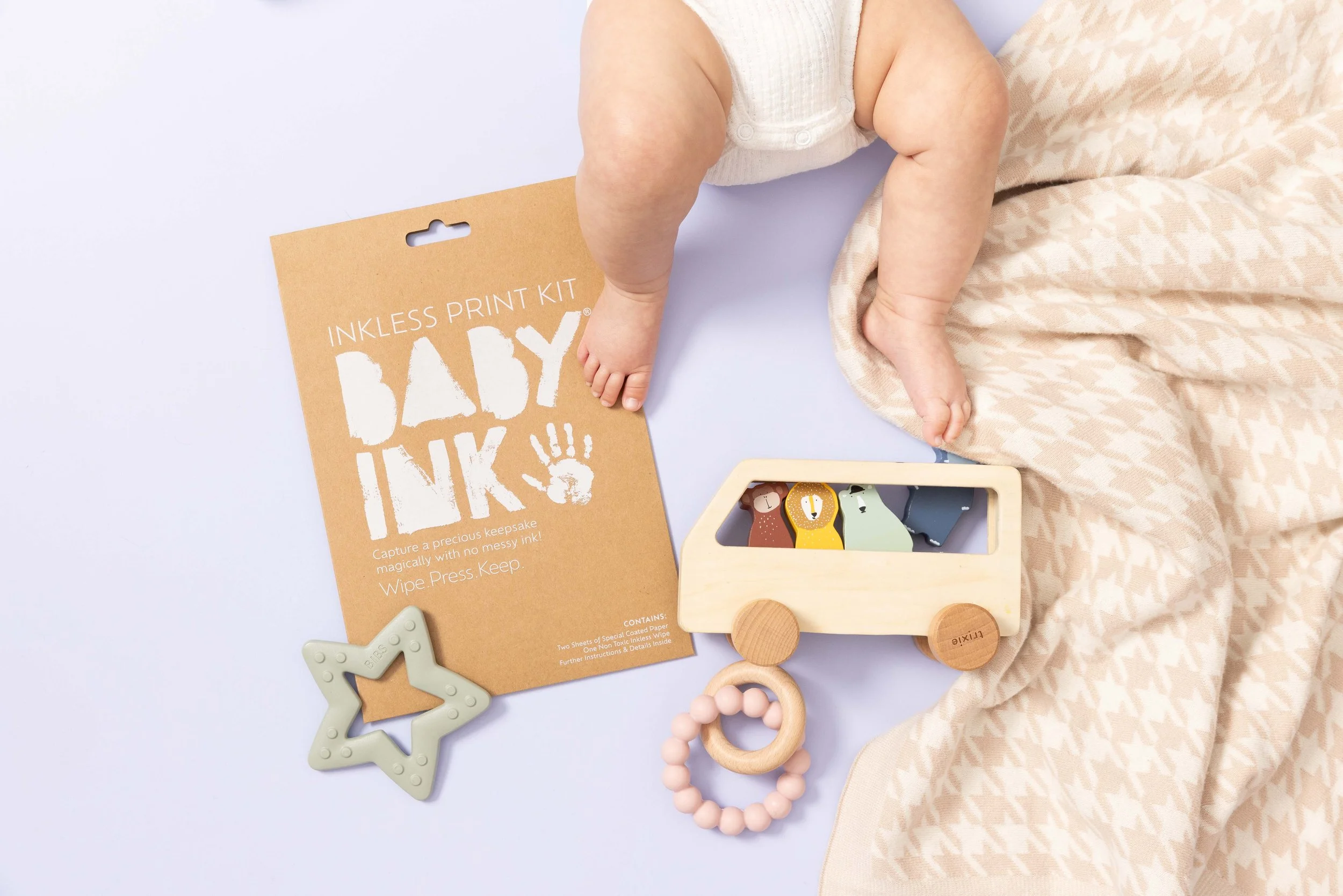 Baby lying on a beige houndstooth blanket next to a wooden toy truck, a pink beaded teething ring, a star-shaped teething toy, and a brown paper package labeled "INKLESS PRINT KIT BABY INK" on a white surface.