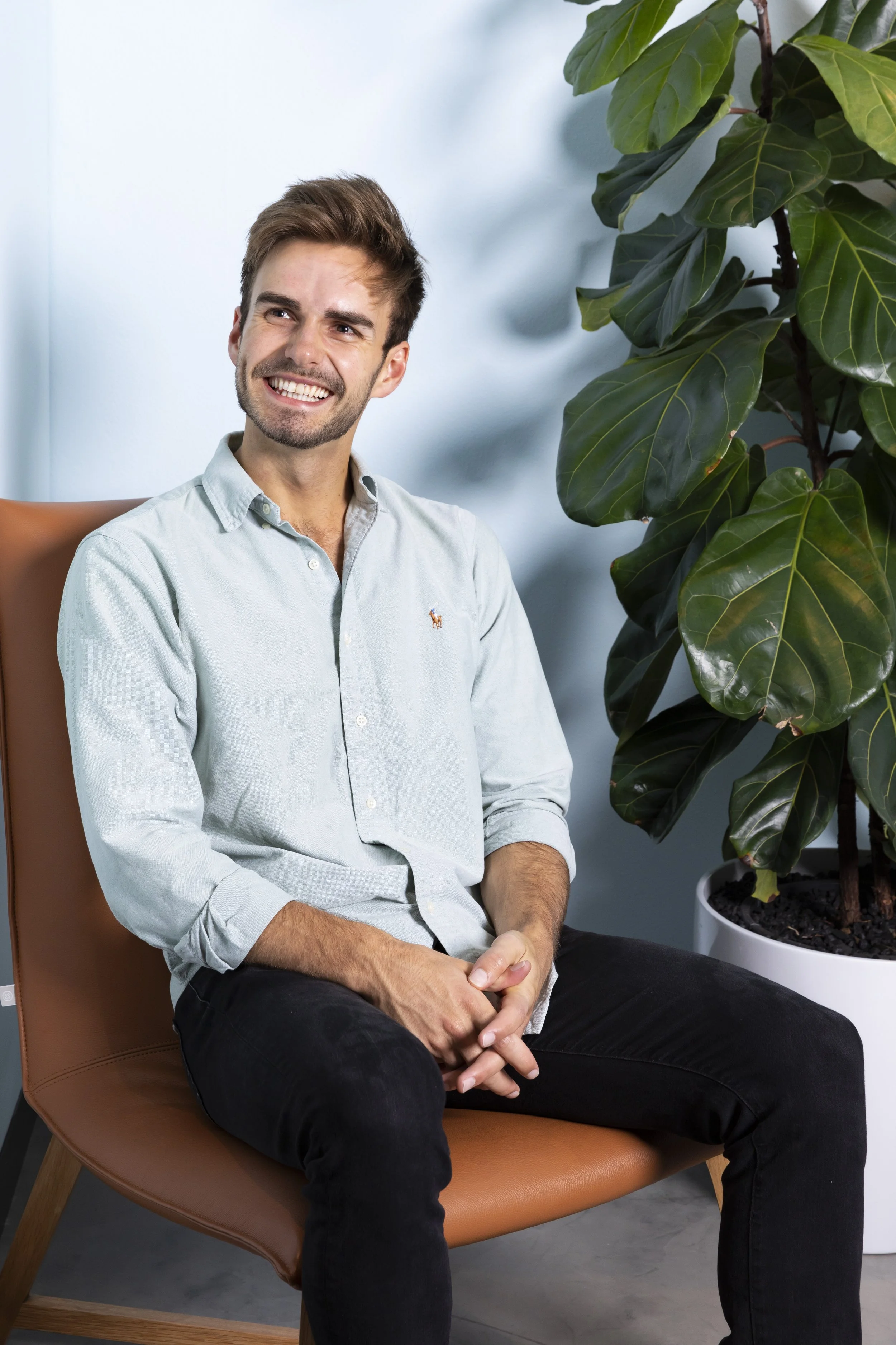 A young man with brown hair and a beard smiling, sitting in a brown chair next to a large potted plant with dark green leaves.