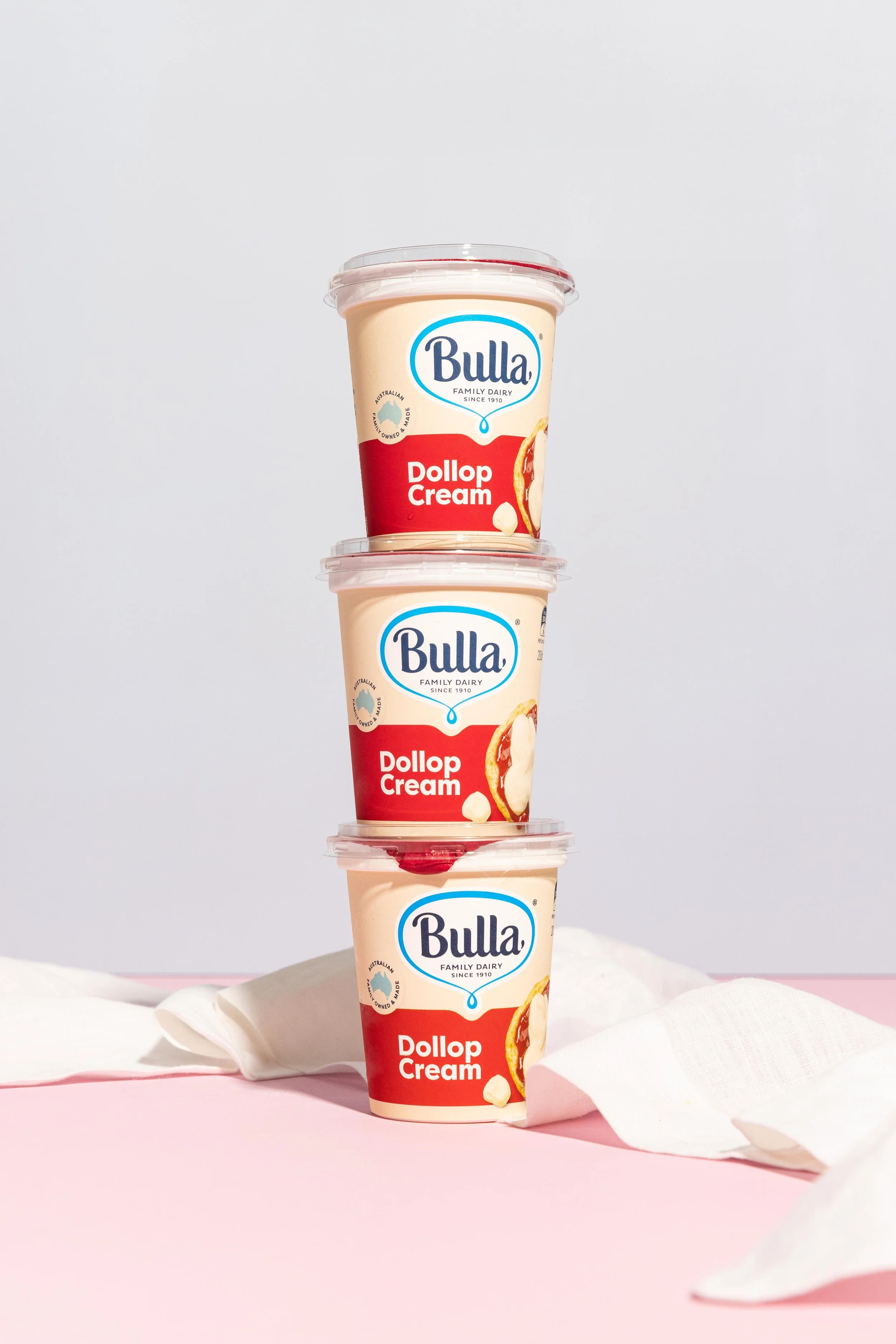 Three stacked cups of Bulla Dollop Cream ice cream with a pink and white background.