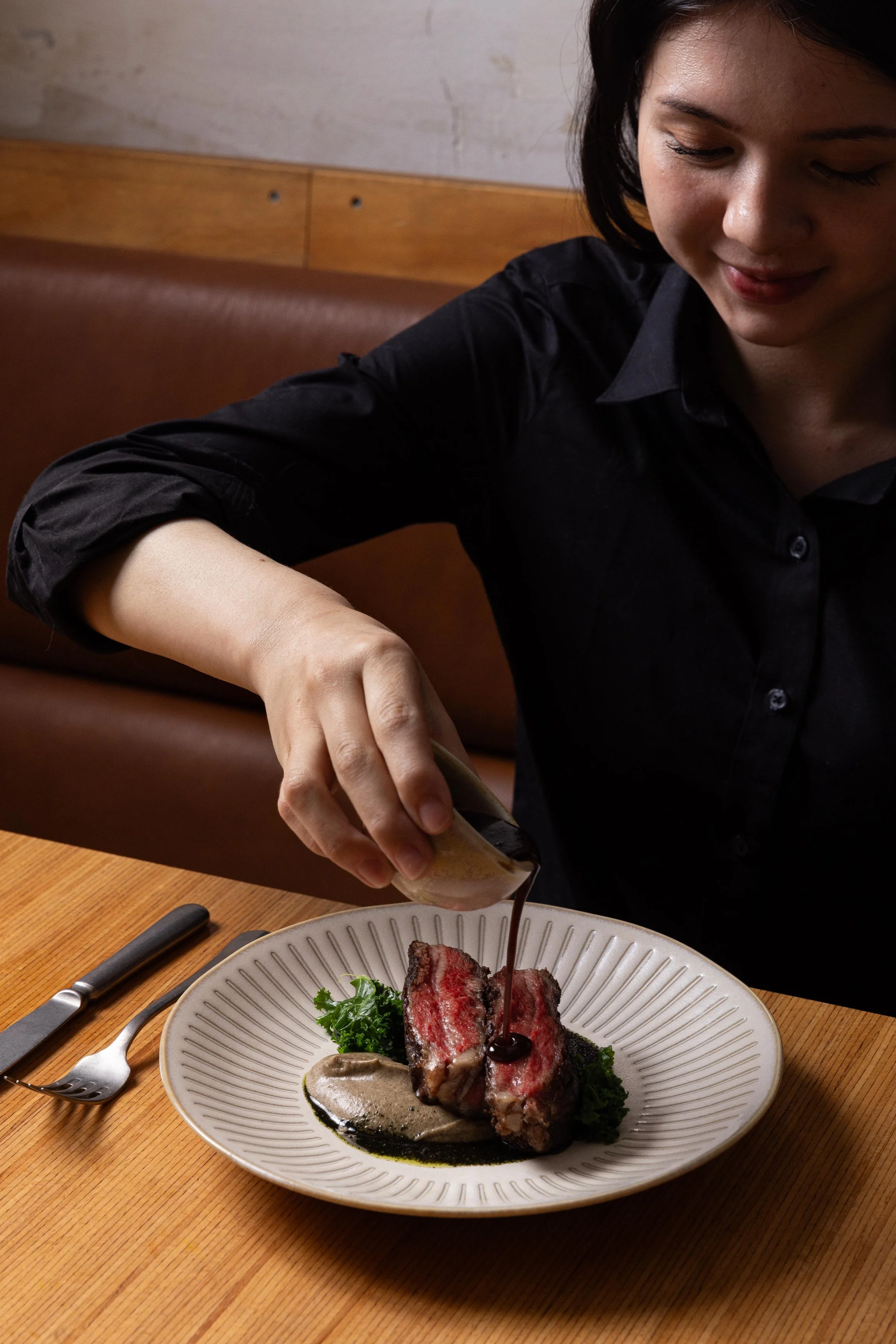 A woman in a black shirt is pouring sauce or gravy onto a piece of cooked steak on a white plate, with greens and a sauce on the plate, in a restaurant setting.